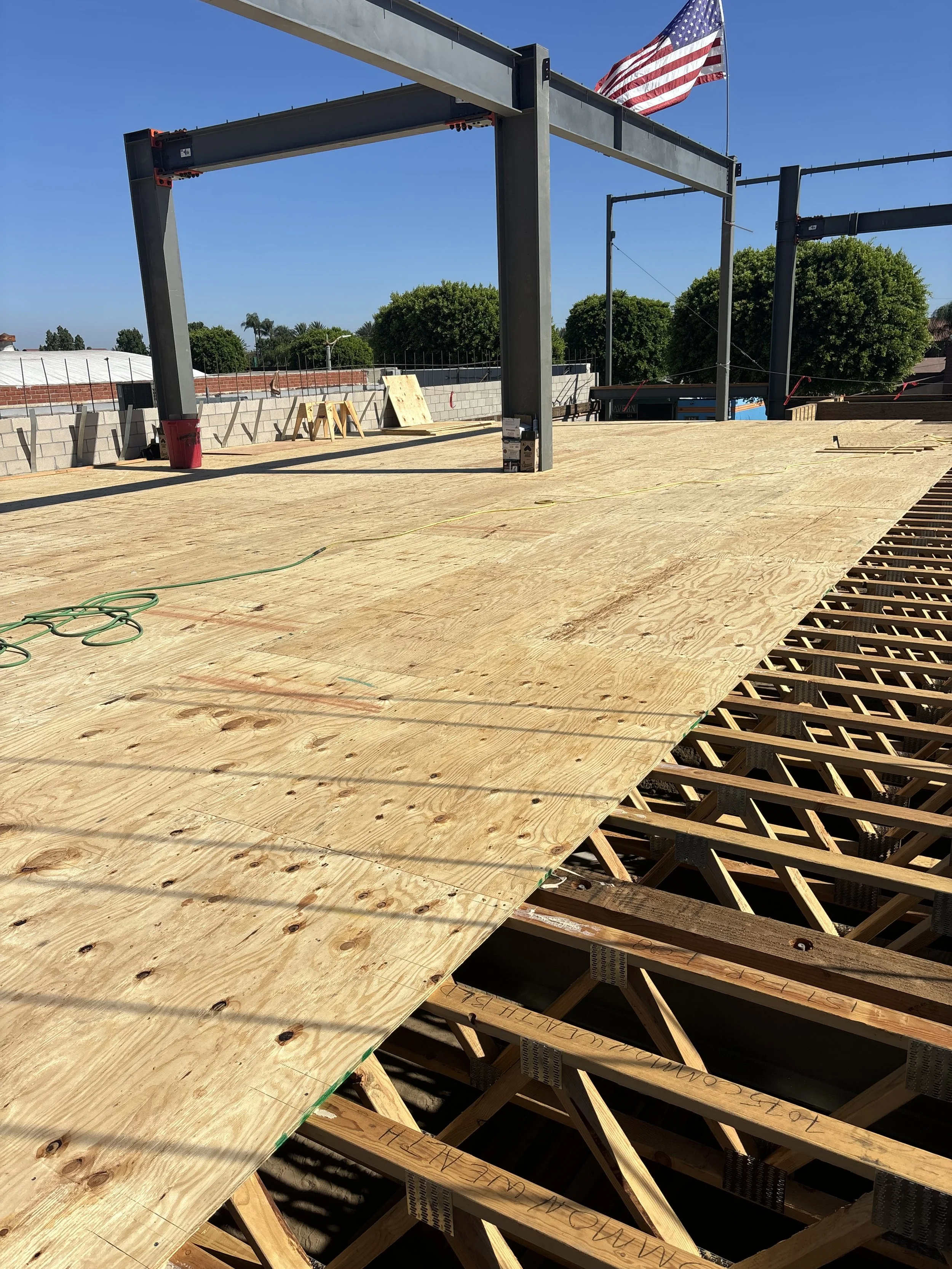 Construction site with wooden floor partially laid, metal framing for a new structure, American flag flying on a pole, and blue sky in the background.