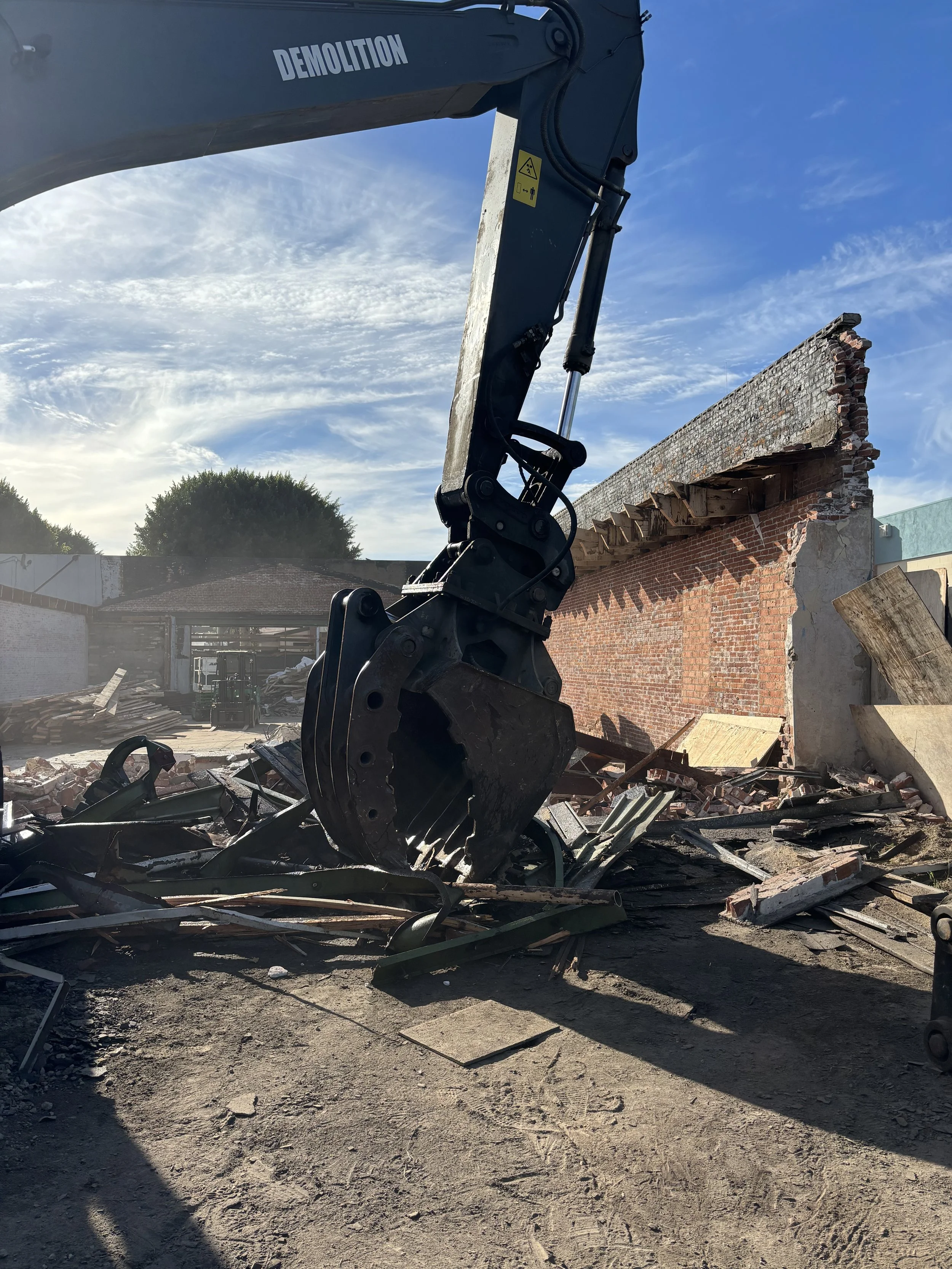 A demolition excavator with a large bucket attachment tearing down a brick wall, debris scattered on the ground under a blue sky with clouds.