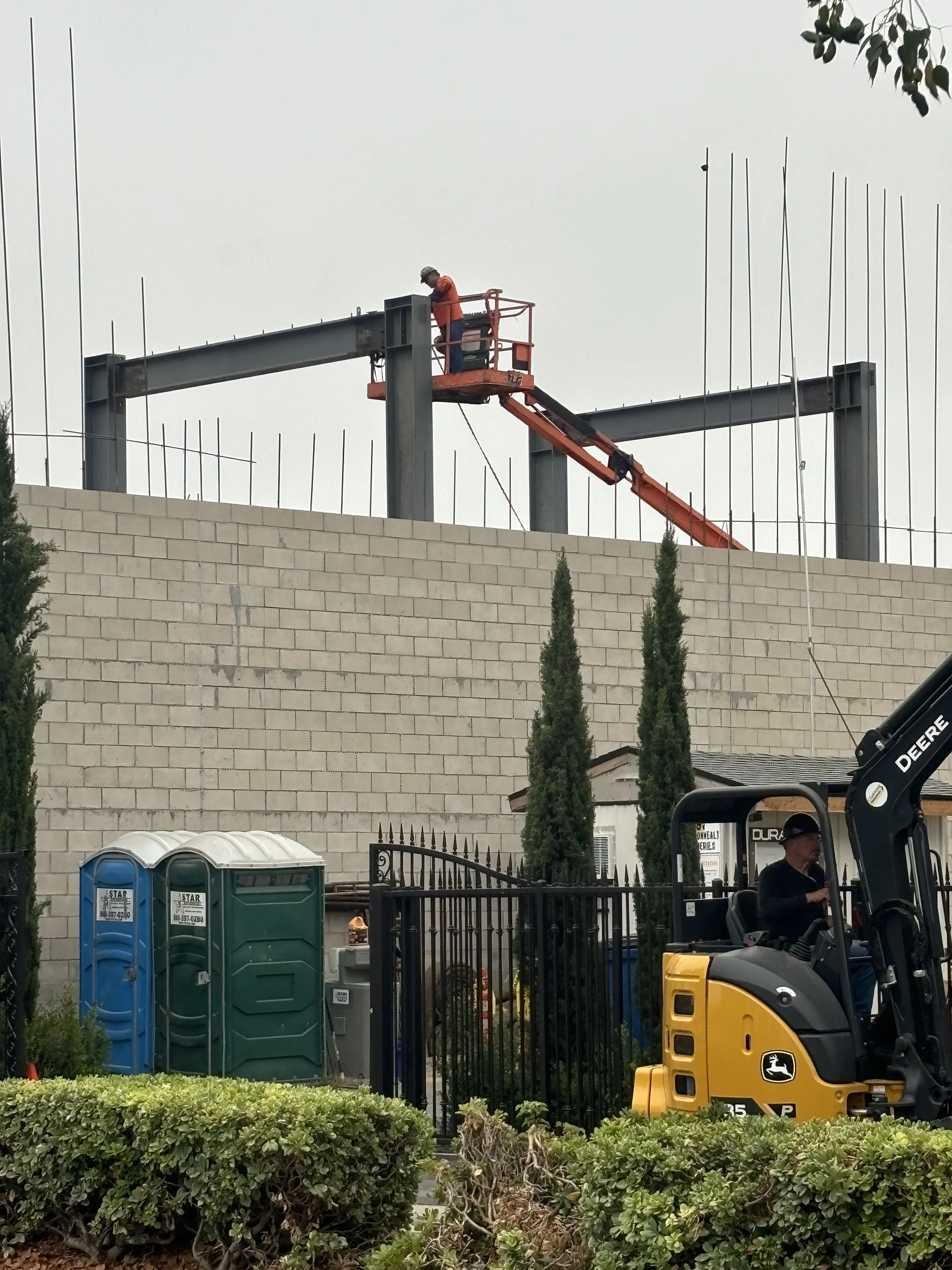 Construction worker standing on orange lift platform working on steel framework above a concrete block wall. Portable toilets, trees, and construction equipment visible below.