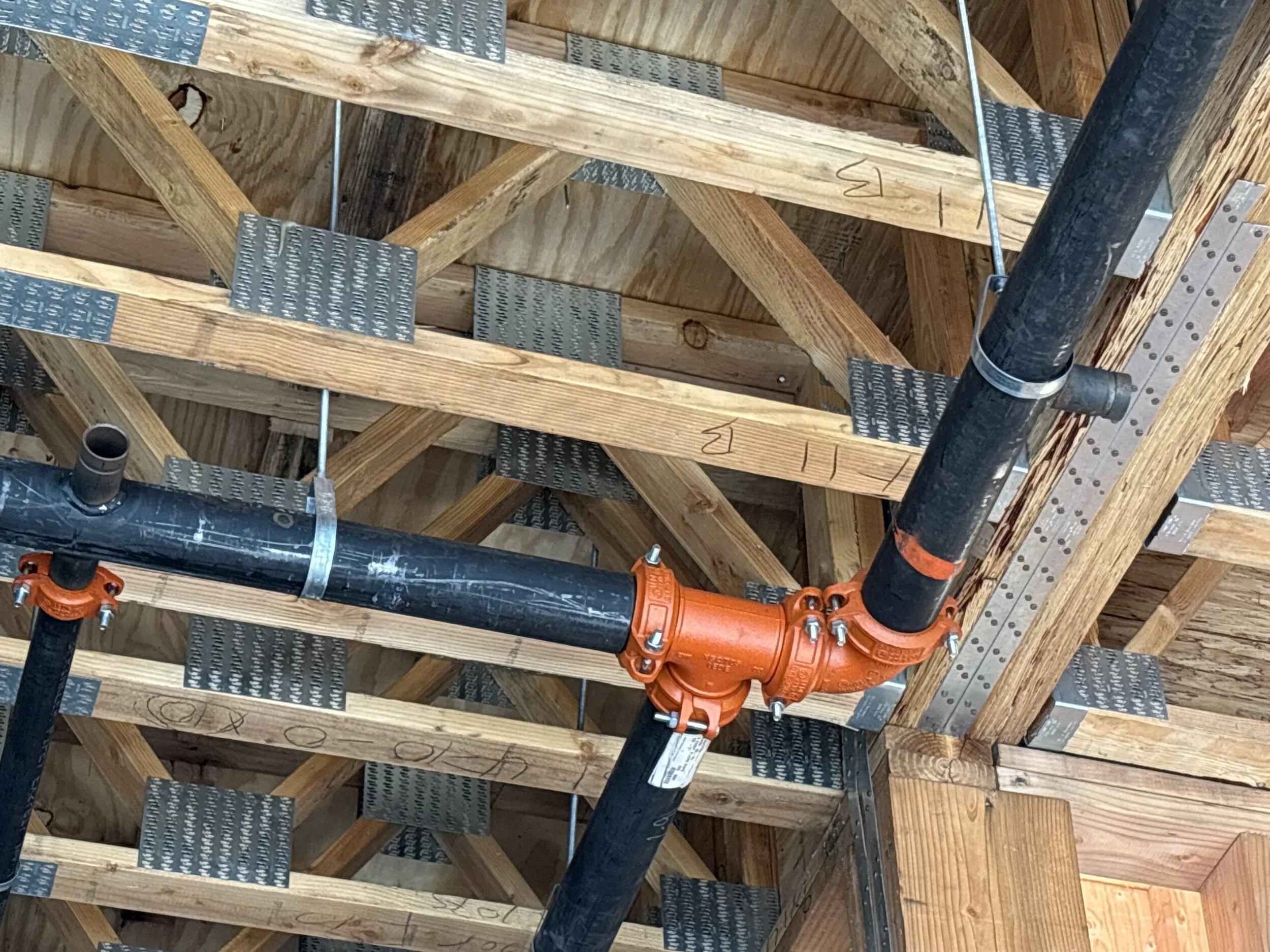View of building construction with wooden beams, metal plates, and black PVC pipes connected with orange fittings.