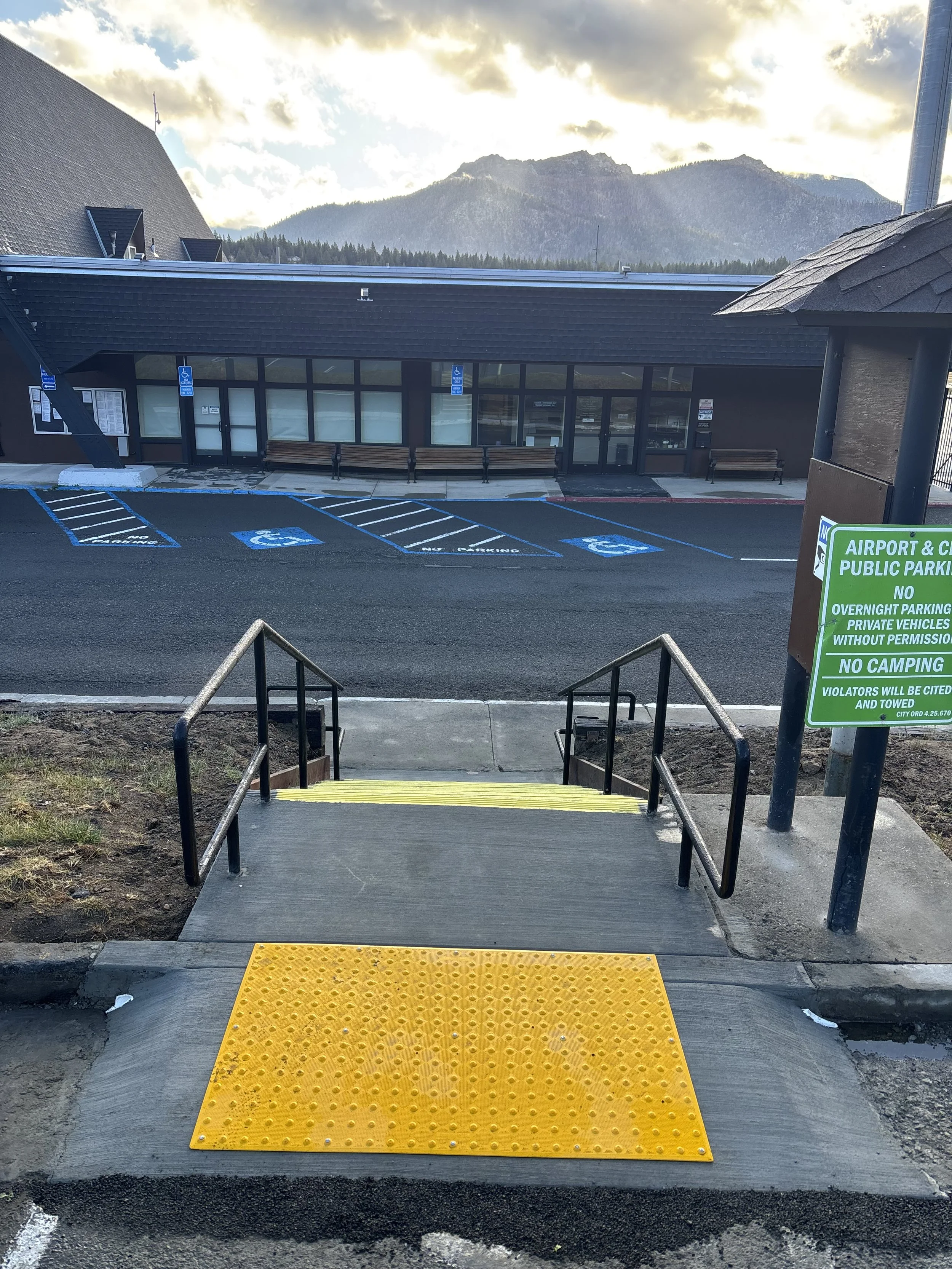 Sidewalk with yellow textured ramp leading down to stairs with handrails, in front of a parking lot with designated handicap spaces and a building with a windowed front, mountains, and cloudy sky in the background.