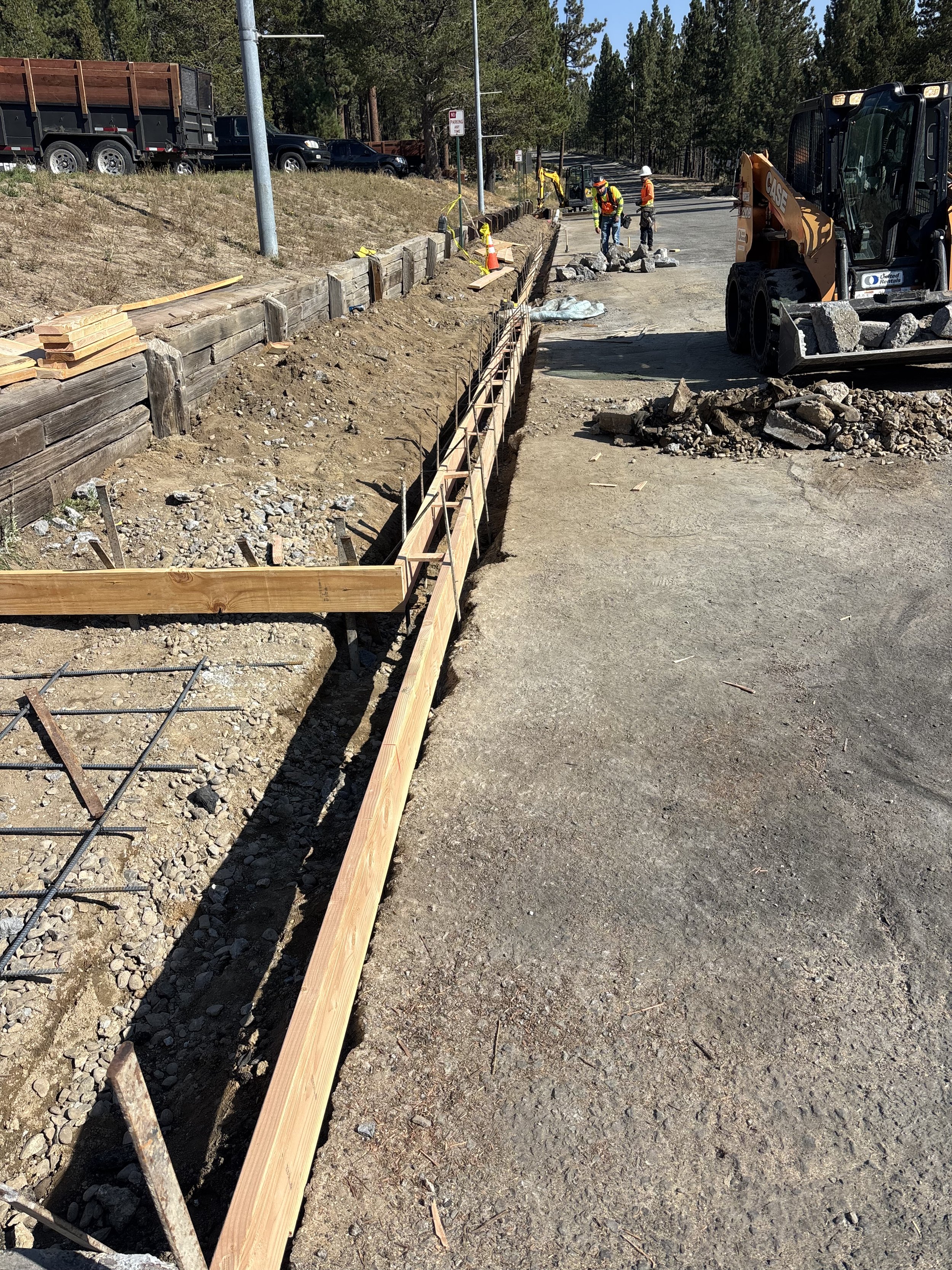 Construction workers and machinery working on a road expansion project with wooden forms and dirt.