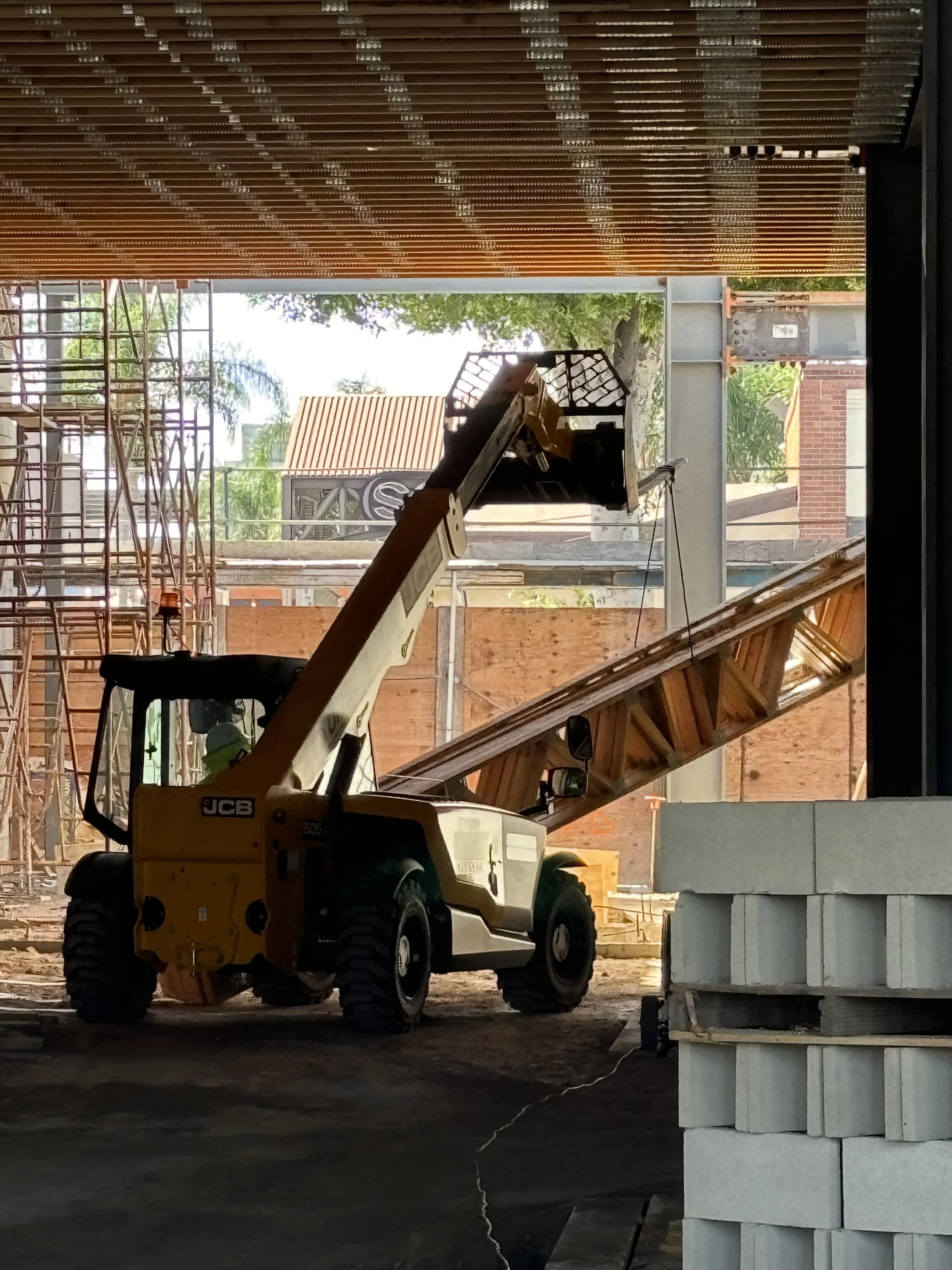 Construction site with a yellow JCB telescopic forklift lifting materials, scaffolding on the left, and brick and wooden structures in the background.