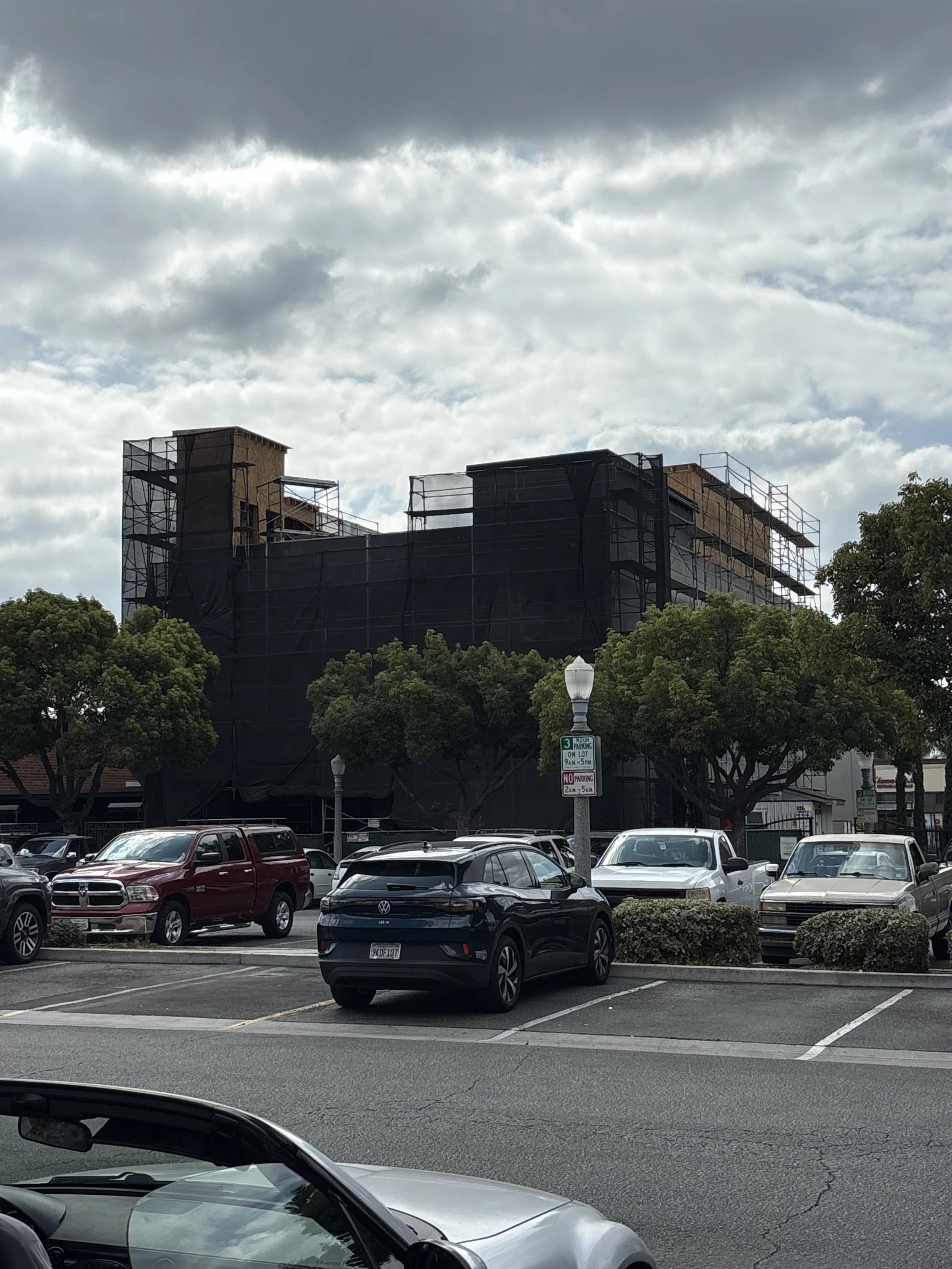 Building under construction covered with black tarp in a parking lot, surrounded by trees and parked cars, with cloudy sky.