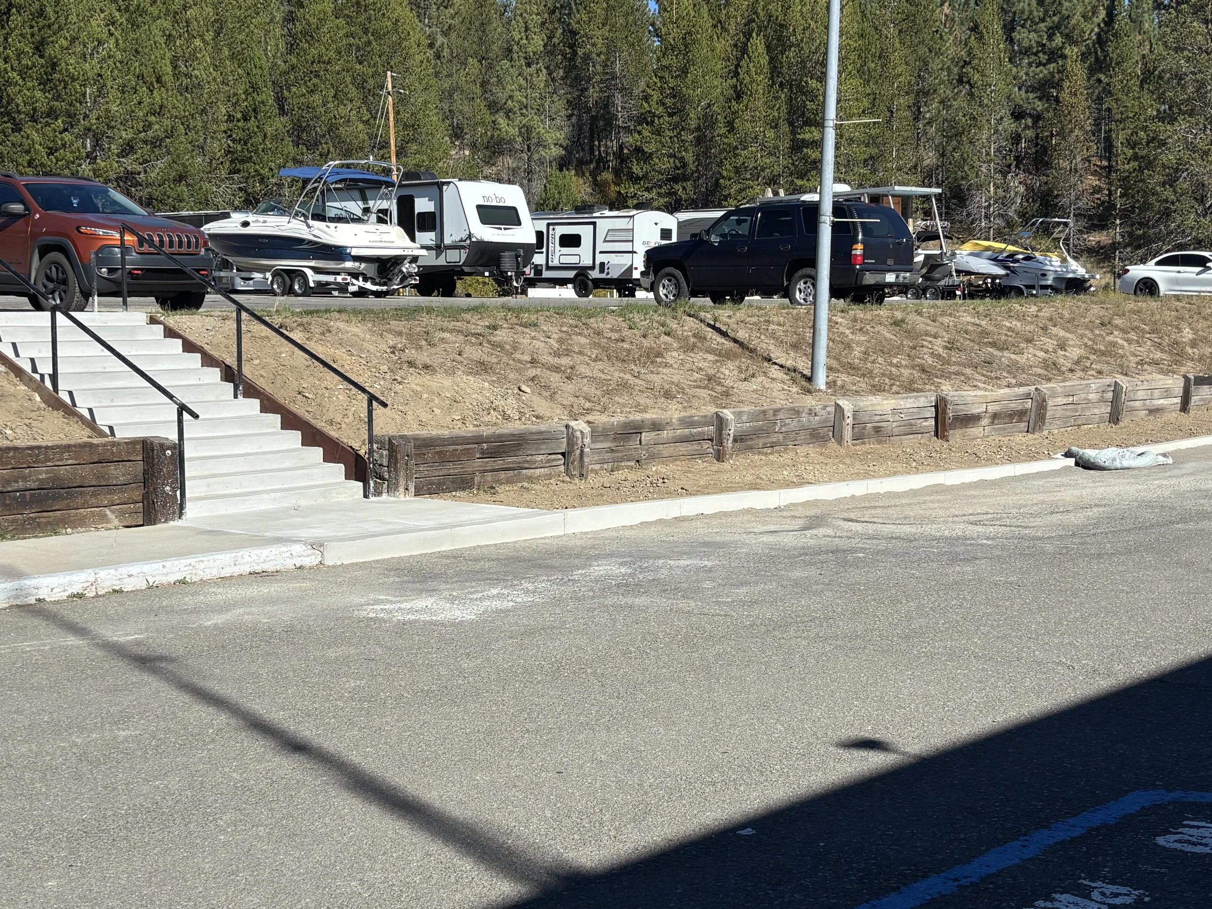 A parking lot with a row of vehicles, including an orange SUV, travel trailers, boats, and several cars, with a background of trees and a clear sky.
