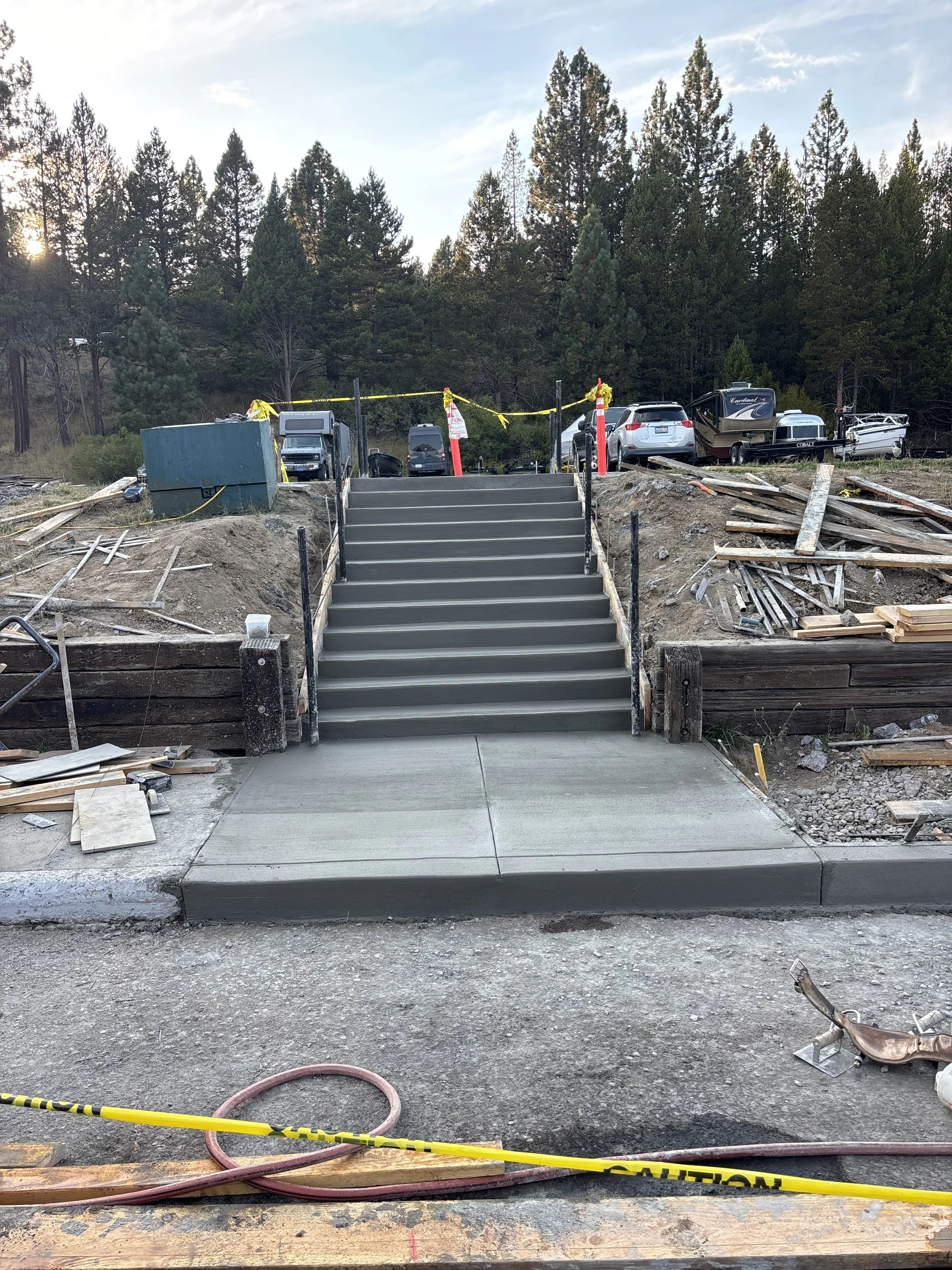 Newly constructed concrete sidewalk and stairs at a construction site with parked cars and trucks in the background.