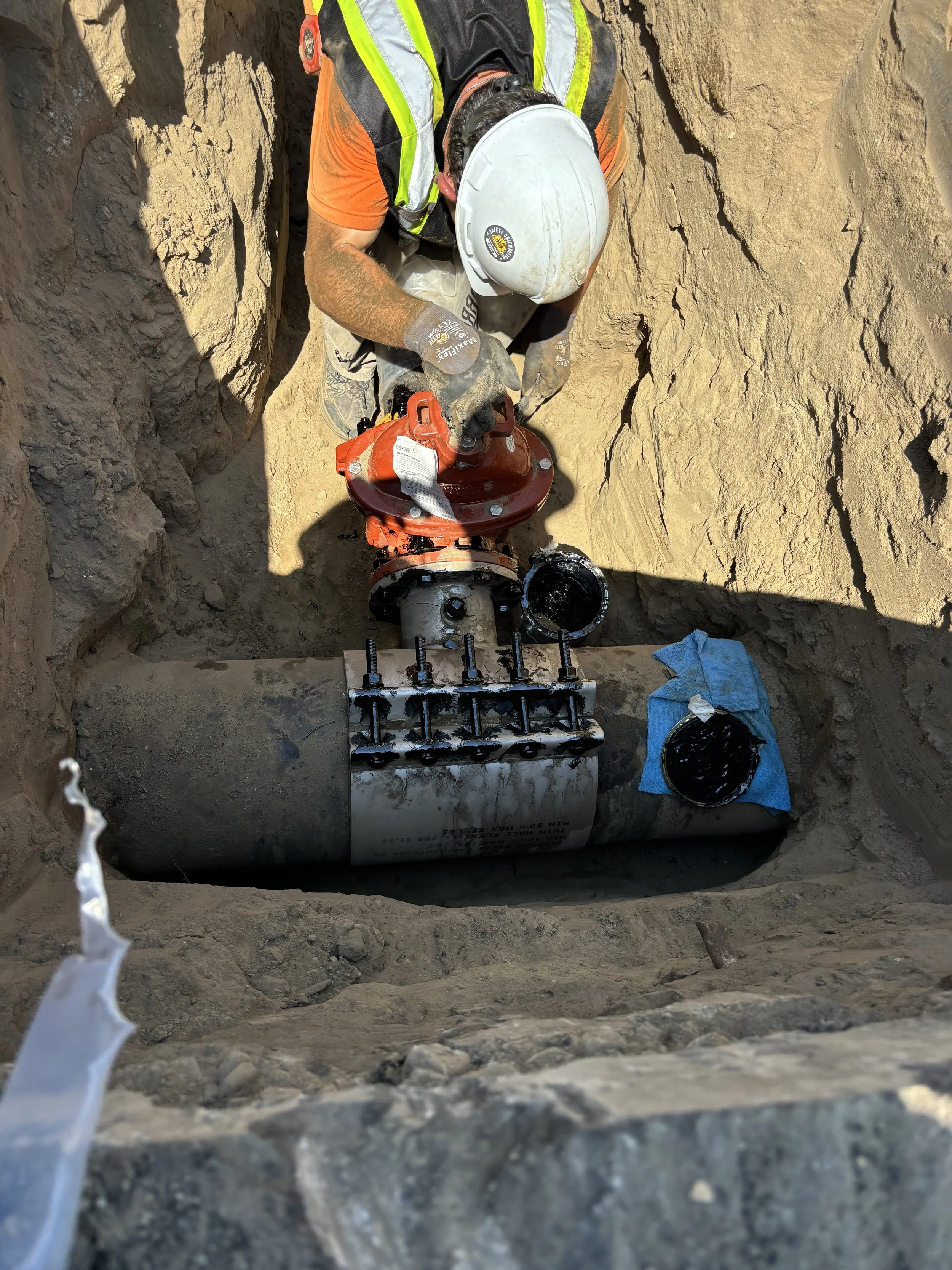 Worker installing or repairing underground pipeline with large valve and pipe fittings, wearing safety gear including a hard hat and gloves.