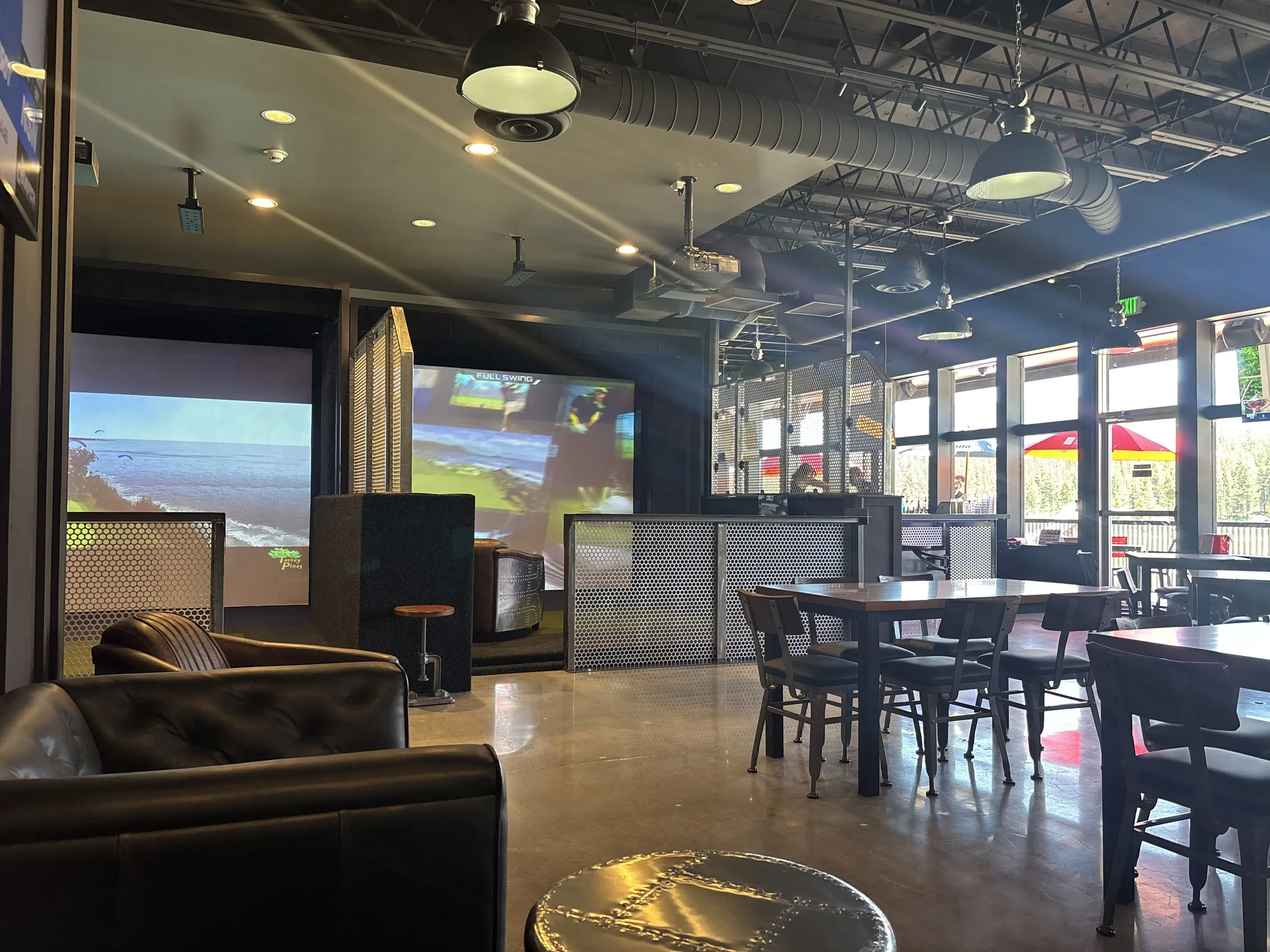 Interior of a sports bar with tables, chairs, large TV screens showing a golf game, and large windows allowing natural light.