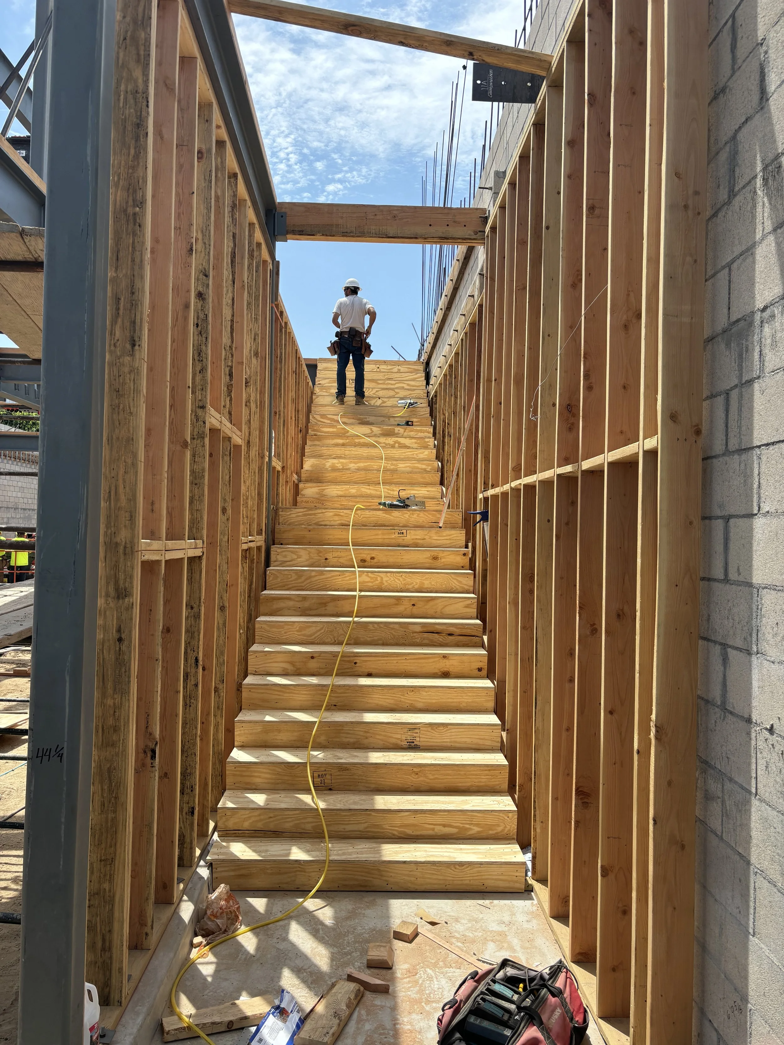 Construction worker standing at the top of a wooden staircase under construction, with framed walls and a blue sky with clouds in the background.