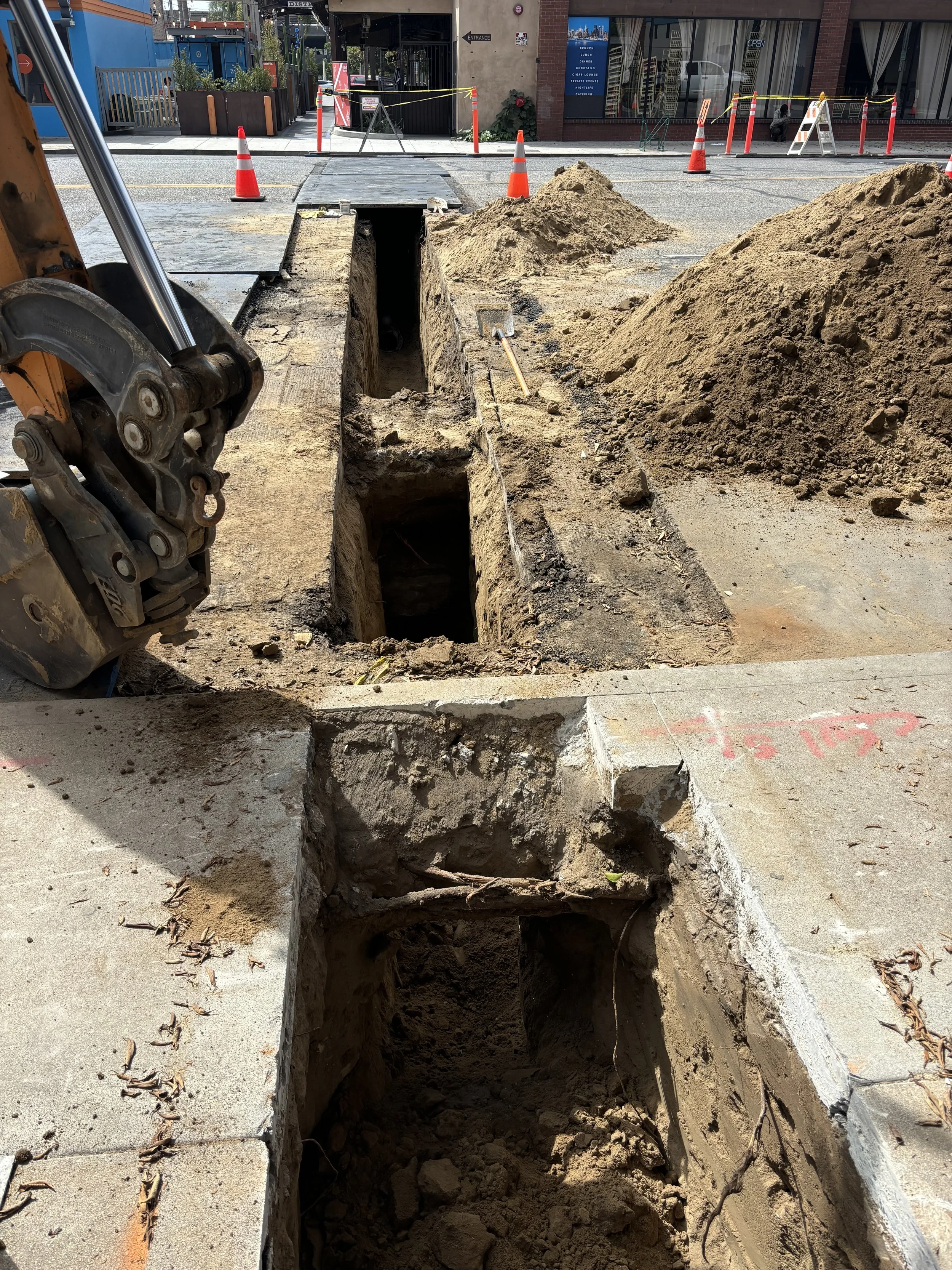 Construction site with a trench dug in the sidewalk, orange safety cones, large piles of dirt, and an excavator arm on the left. Buildings and barriers are visible in the background.
