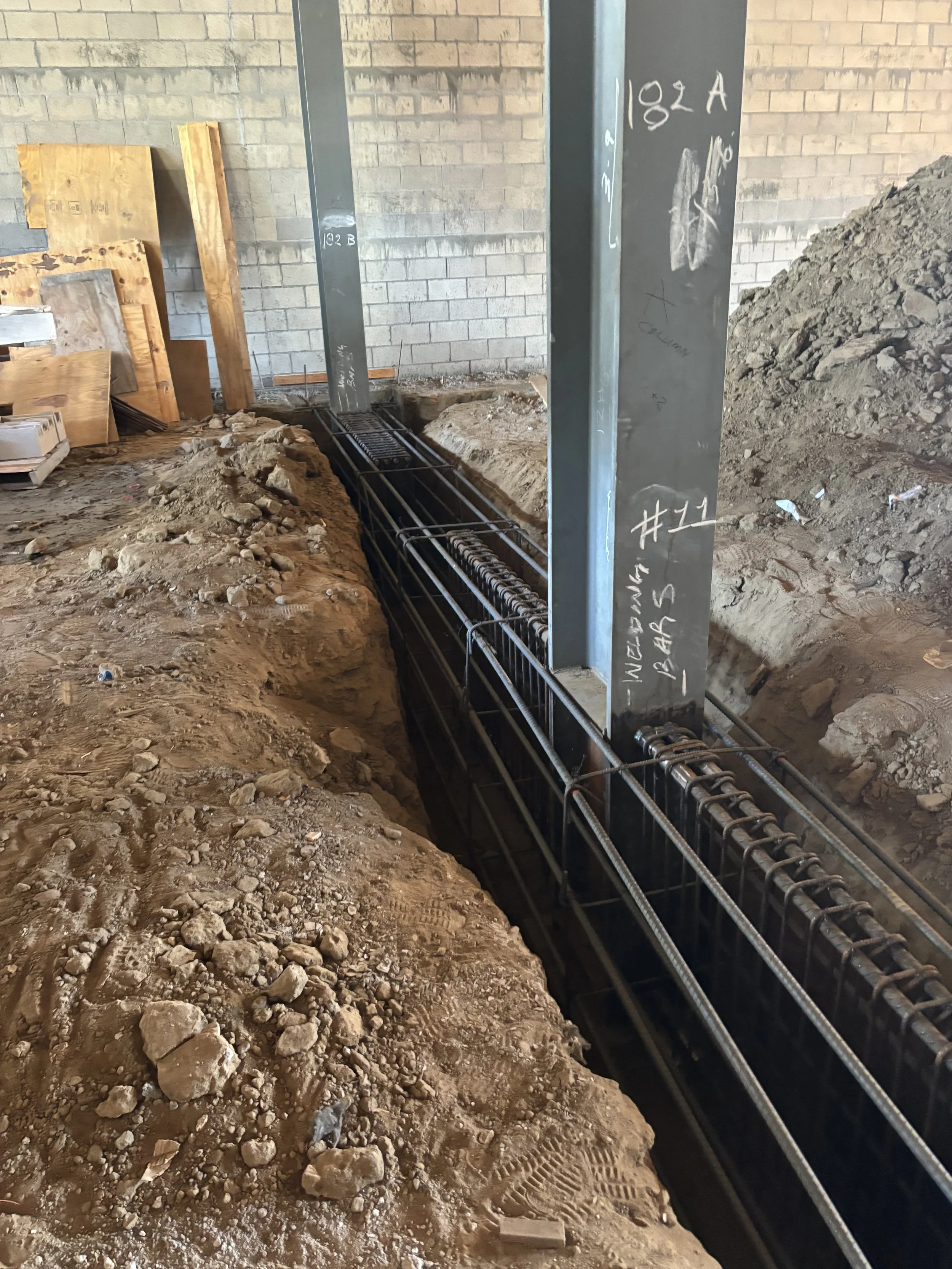 Construction site with trenches filled with reinforcement bars and steel supports for a concrete structure, dirt and debris on the ground, unfinished brick walls, and wooden planks leaning against the wall.