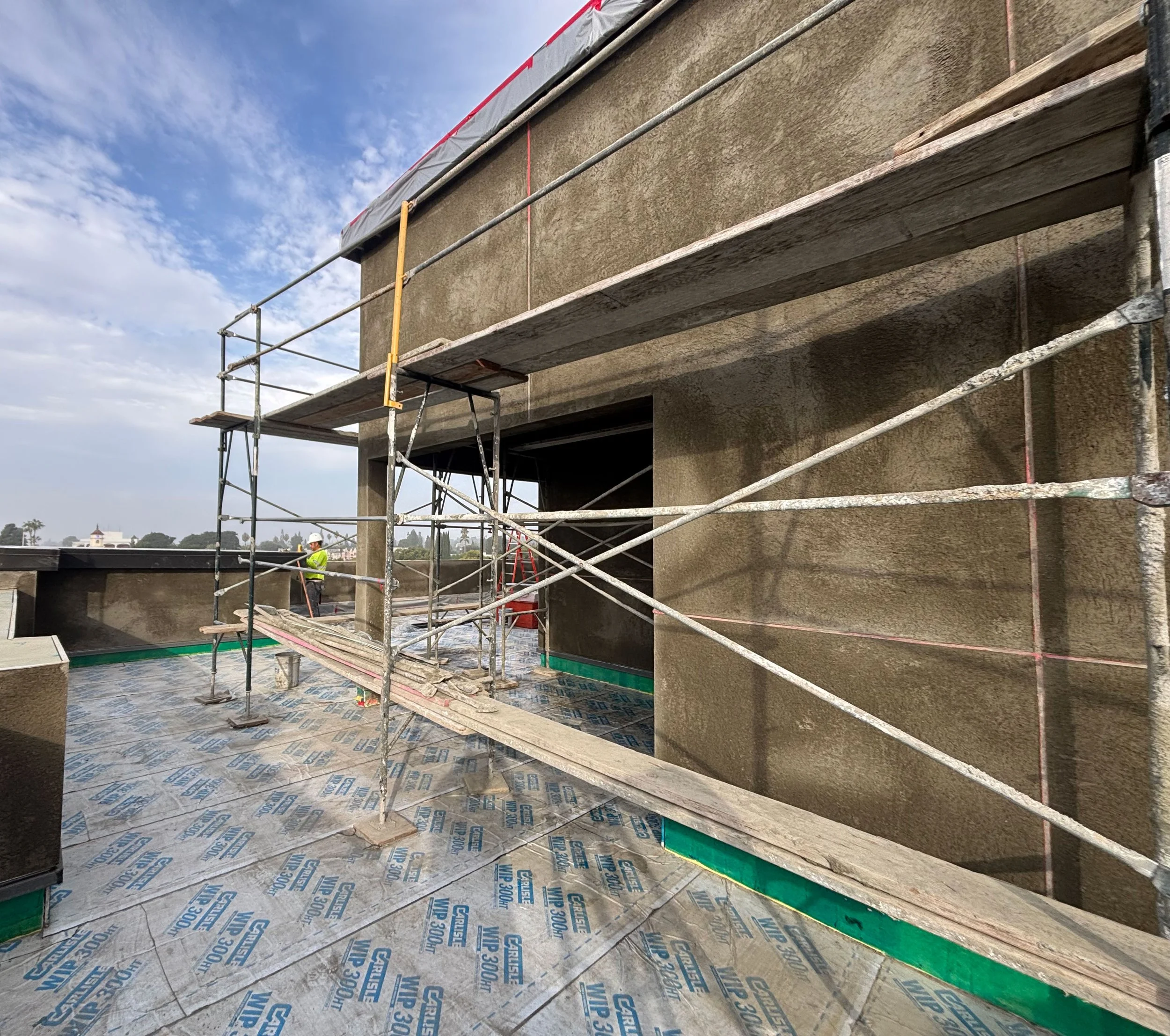 Construction site on a building rooftop with scaffolding and safety equipment, workers in hard hats in the background, and cloudy sky overhead.