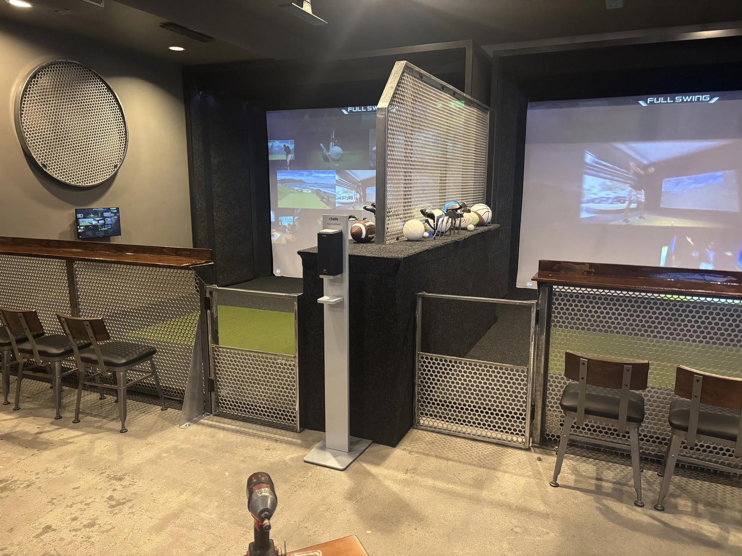 Indoor golf simulator setup with two large projection screens, golf clubs, and golf balls on a shelf, surrounded by chairs and netted barriers.