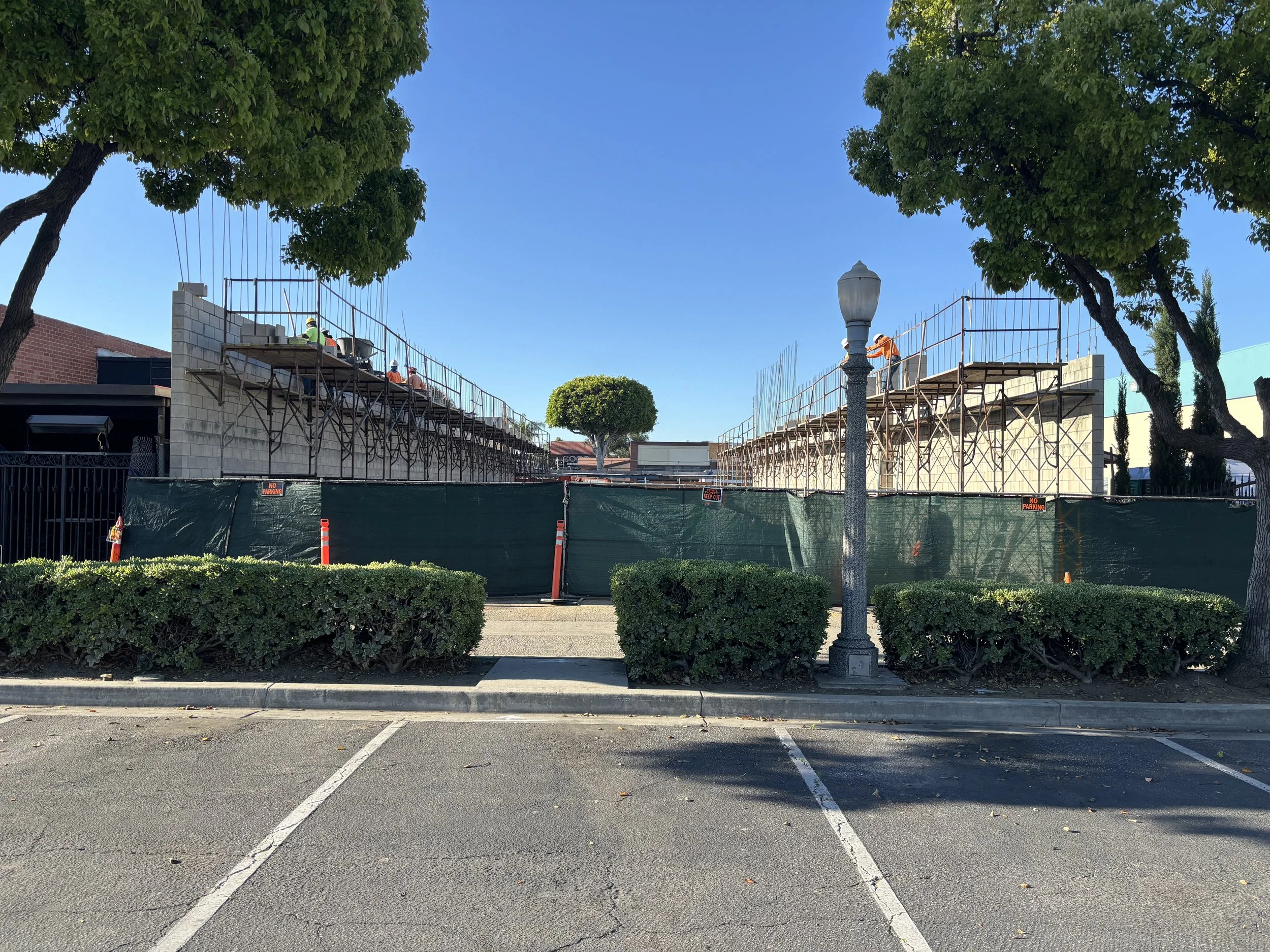 Construction site with scaffolding and workers, surrounded by green fence and bushes, under a clear blue sky.