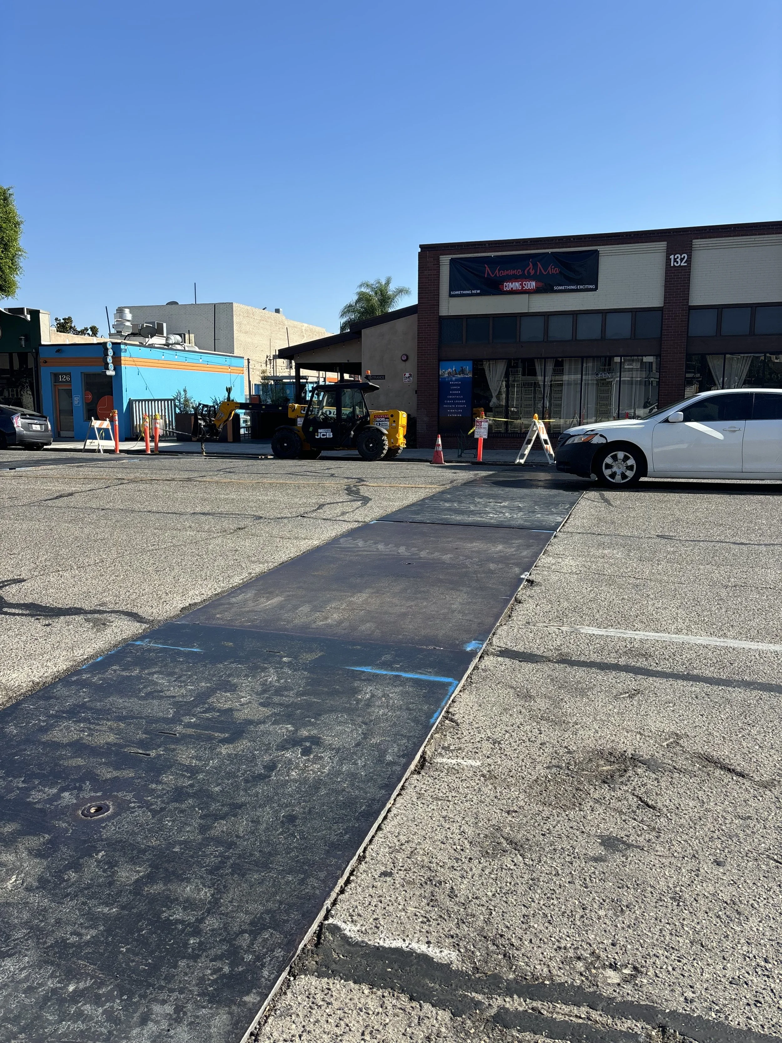 A parking lot with a newly painted handicapped ramp and parking space. There's a white car parked to the right, a yellow construction vehicle in the background, and a building with a banner that reads 'Mamma & Mia Coming Soon.'