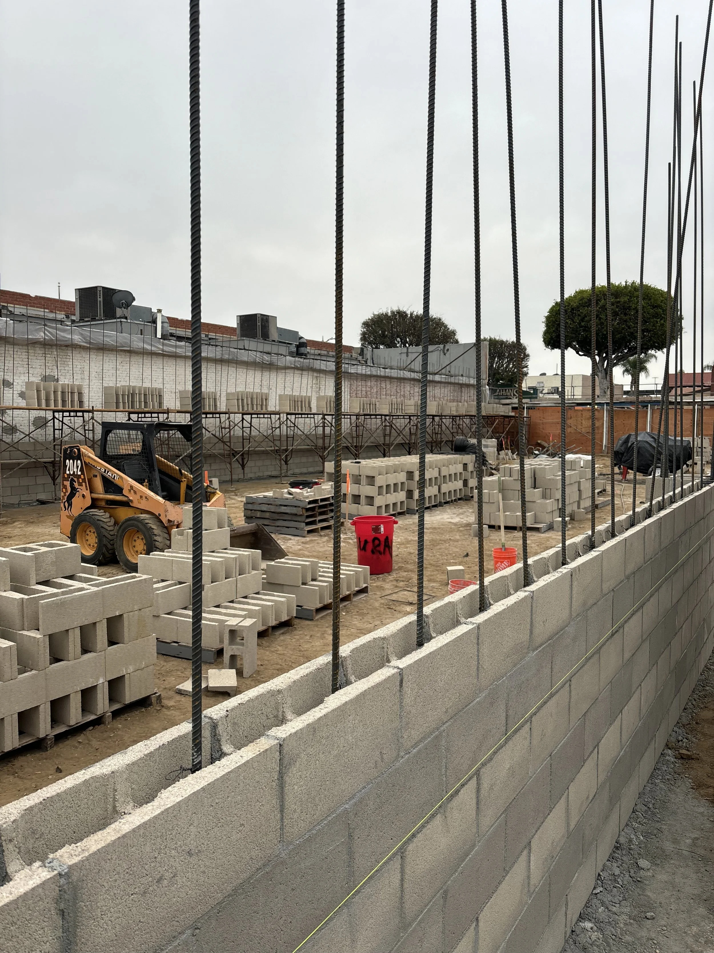 Construction site with concrete blocks, rebar, and construction equipment, enclosed by a wall and situated outdoors with overcast sky.