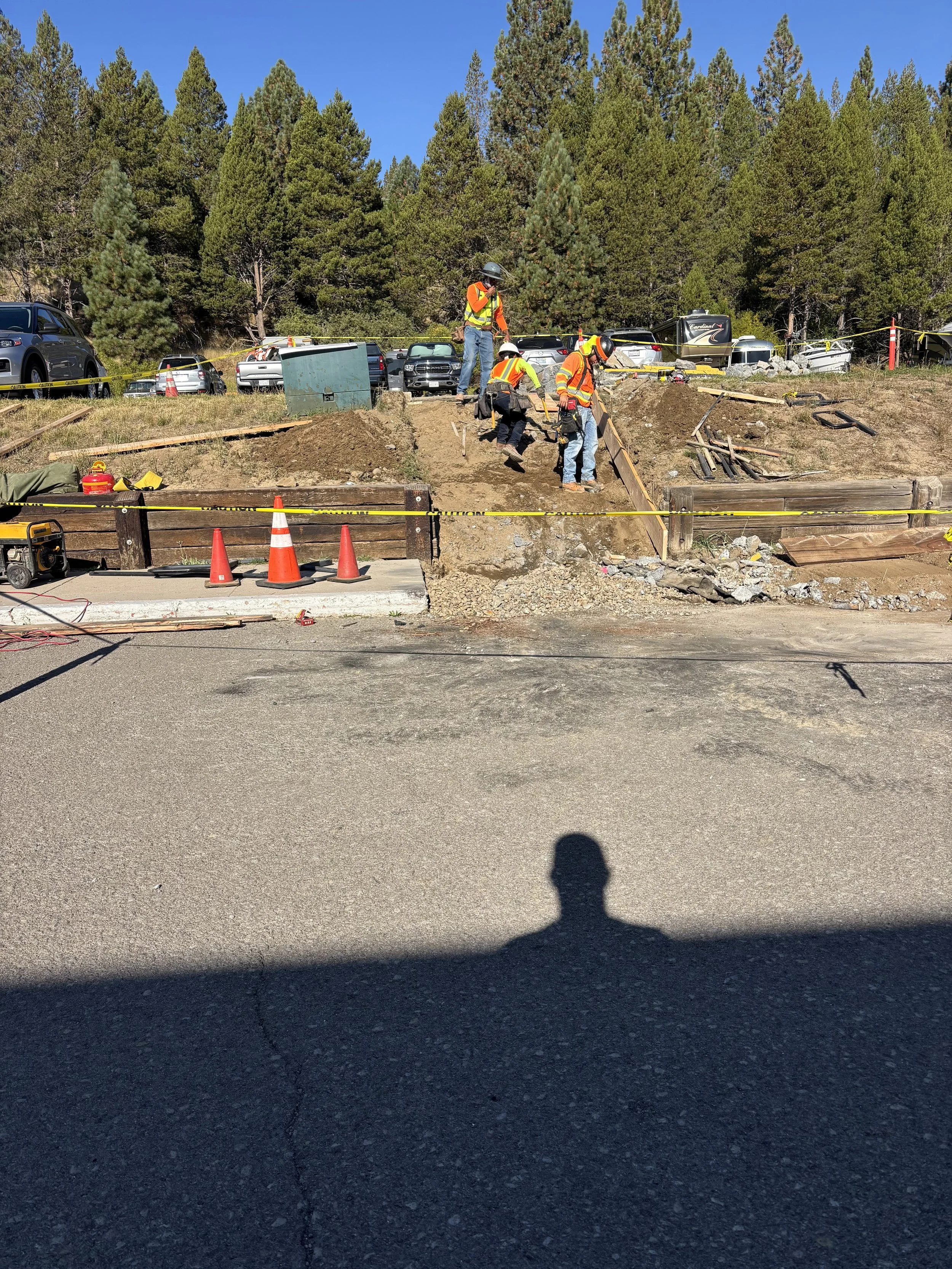Construction workers in safety vests and helmets working on a hillside road construction site with orange traffic cones, parked cars, trees, and a clear blue sky in the background.
