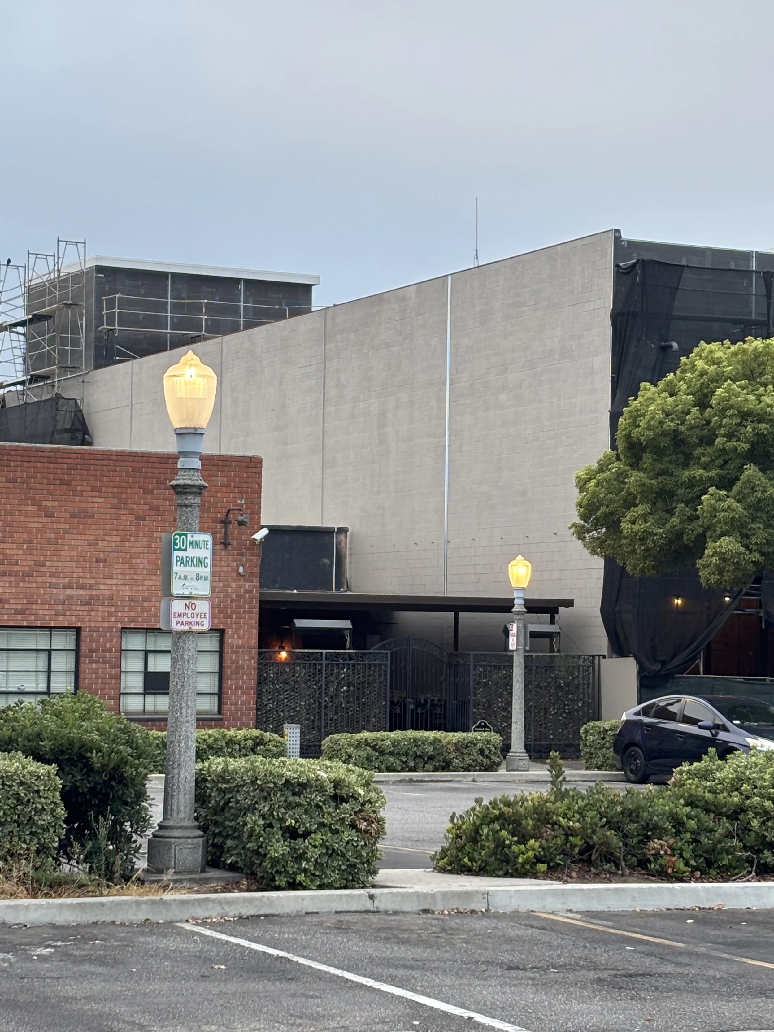 City street scene with a parking lot, two lampposts lit, a black car parked, bushes in the foreground, a brick building, and a large building under construction with scaffolding and black construction netting.