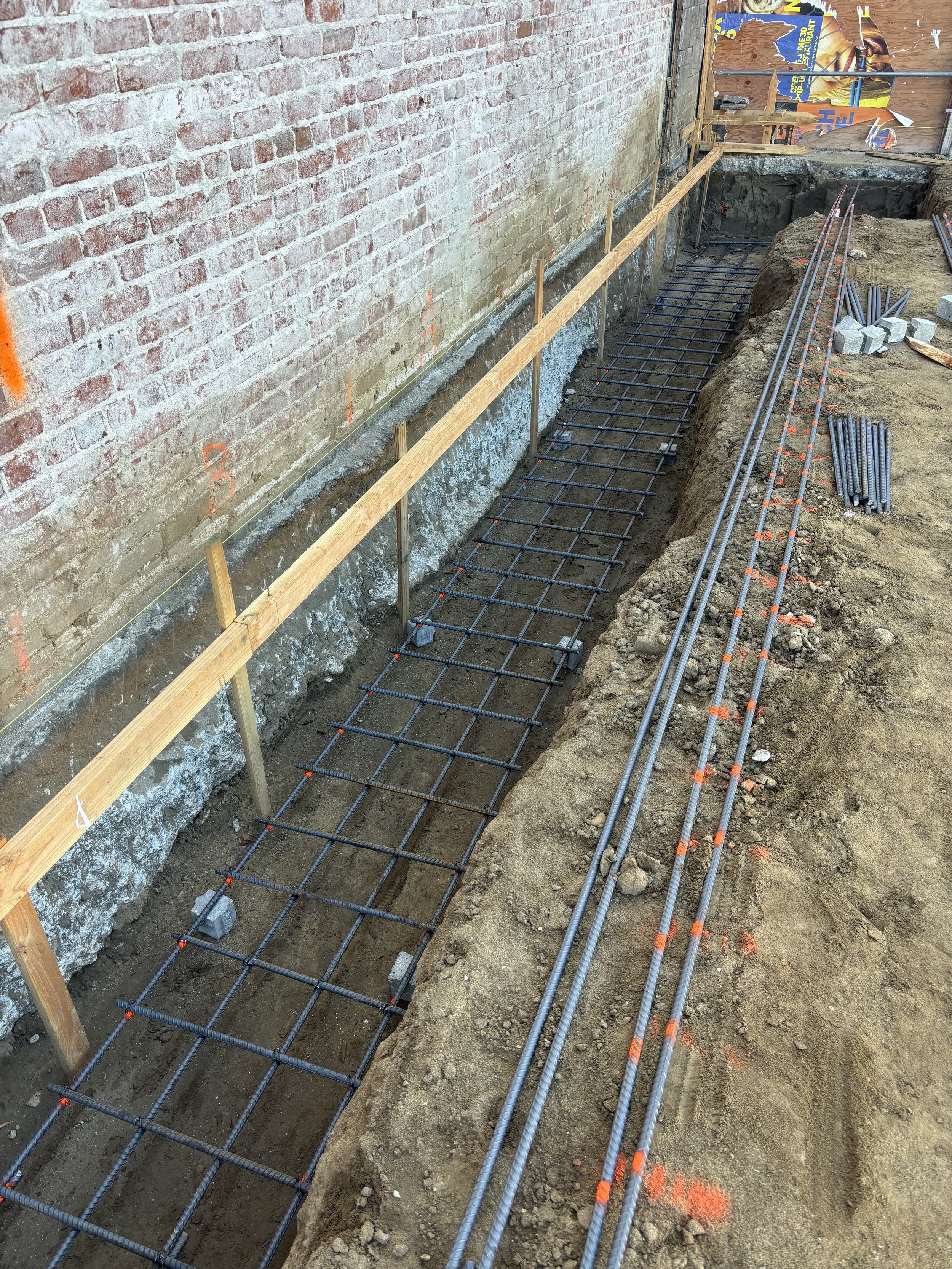 Construction site with rebar grid and wooden formwork for concrete walkway next to a brick wall.