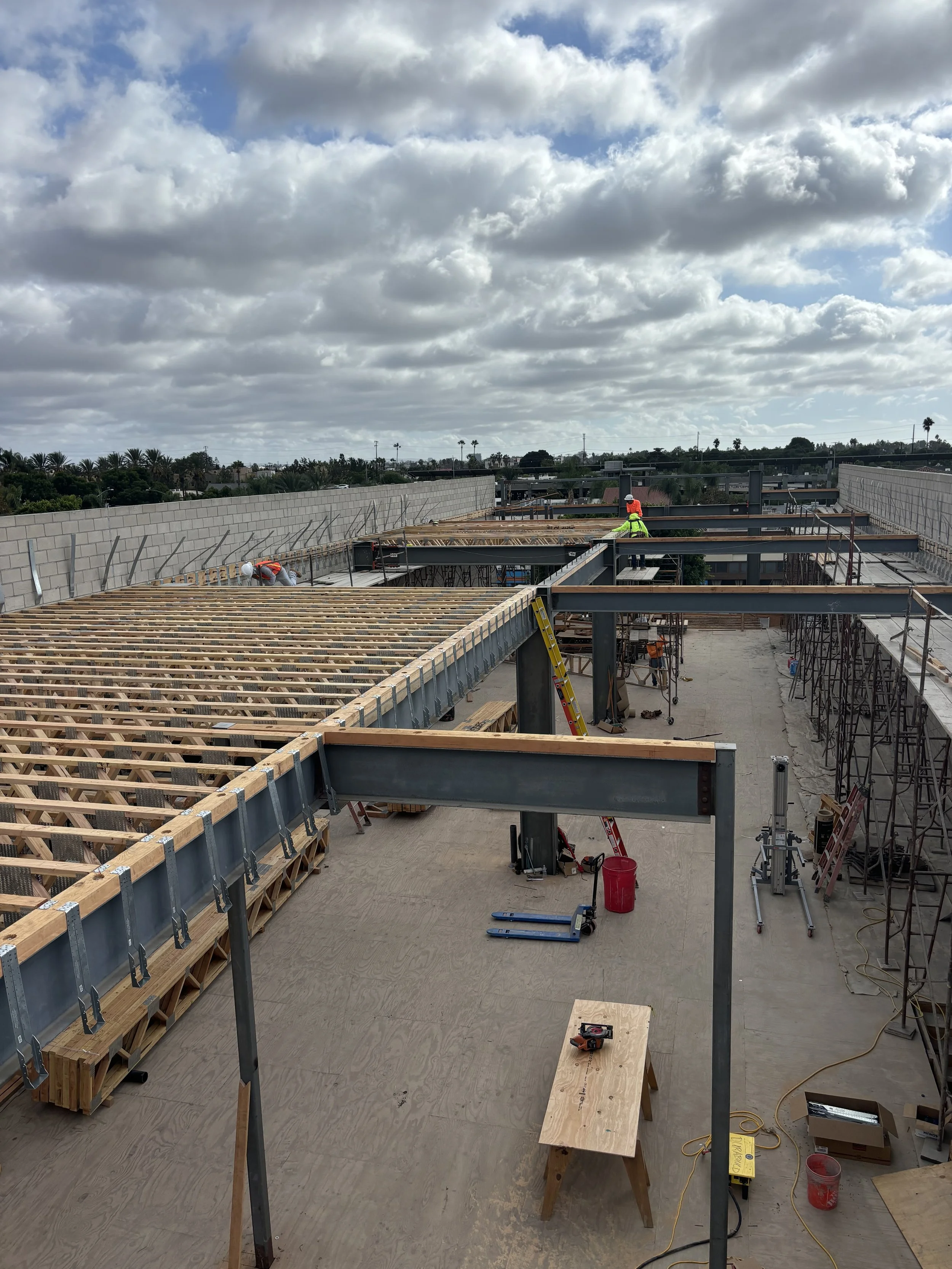 Construction workers on a roof frame under a cloudy sky, with steel beams and wooden planks in progress.