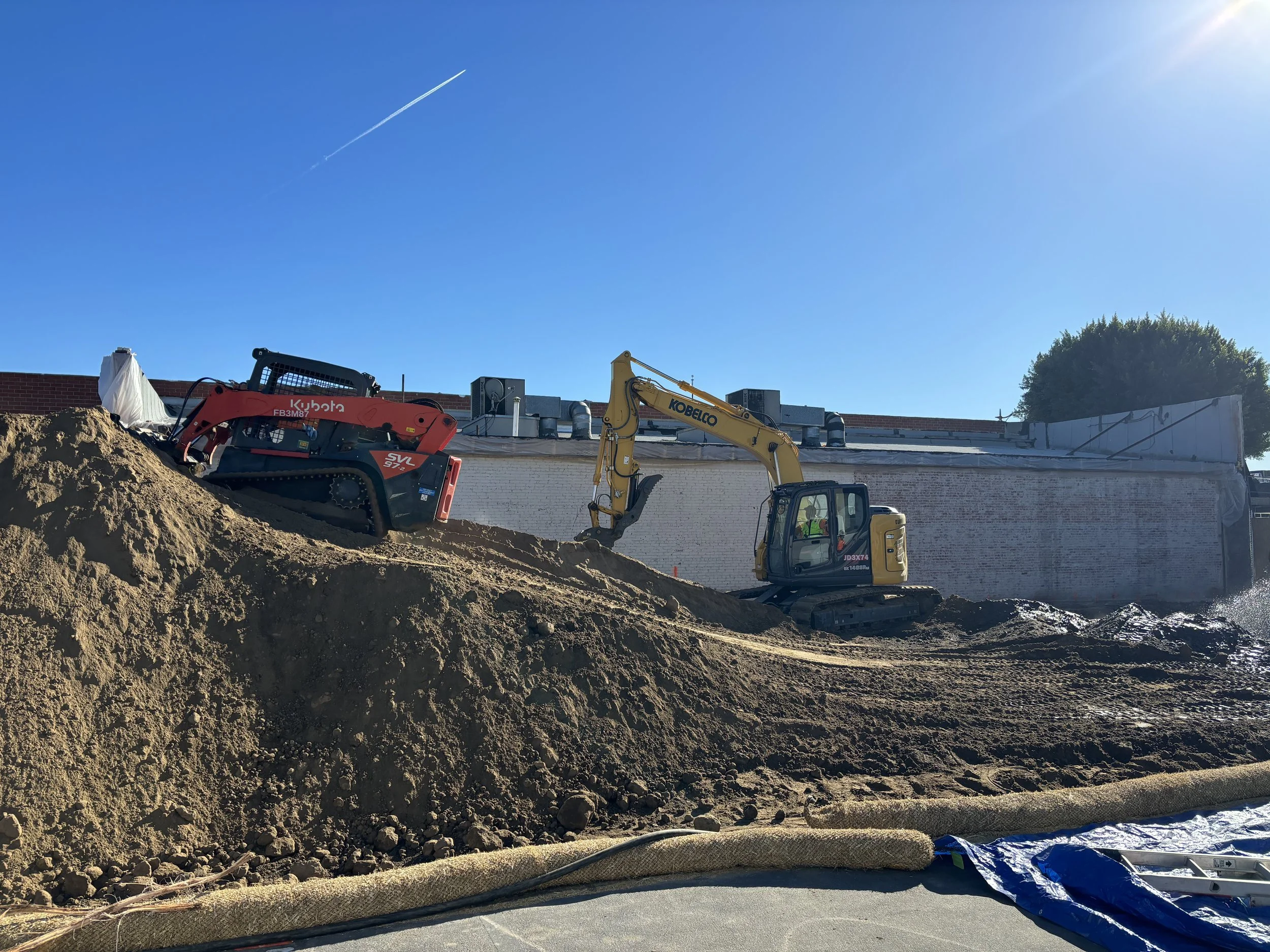 Construction site with a yellow excavator and a red compact loader working on a dirt mound under clear blue sky.