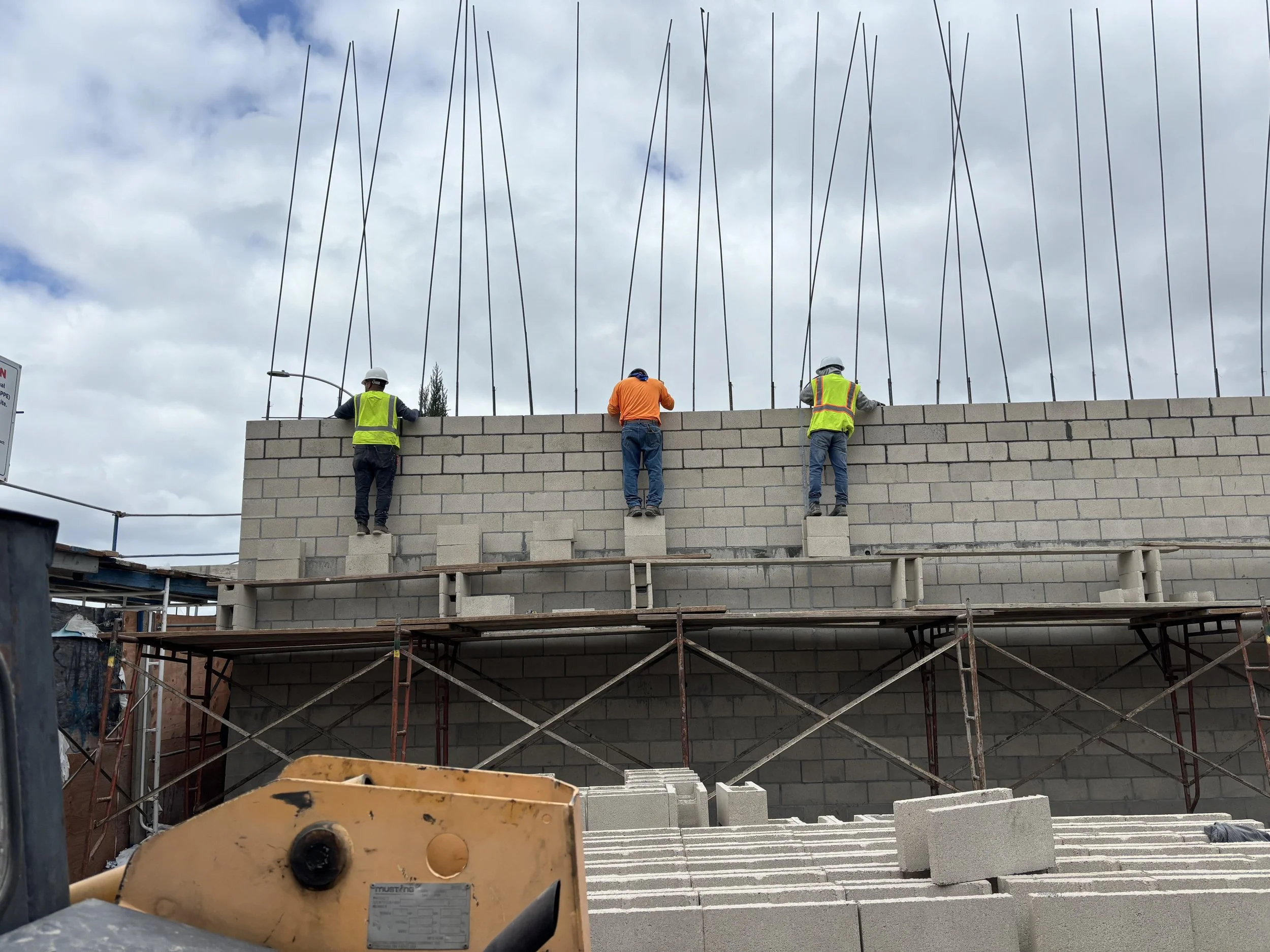 Three construction workers in safety vests and helmets working on a brick wall at a construction site, with scaffolding and building materials below and a cloudy sky overhead.