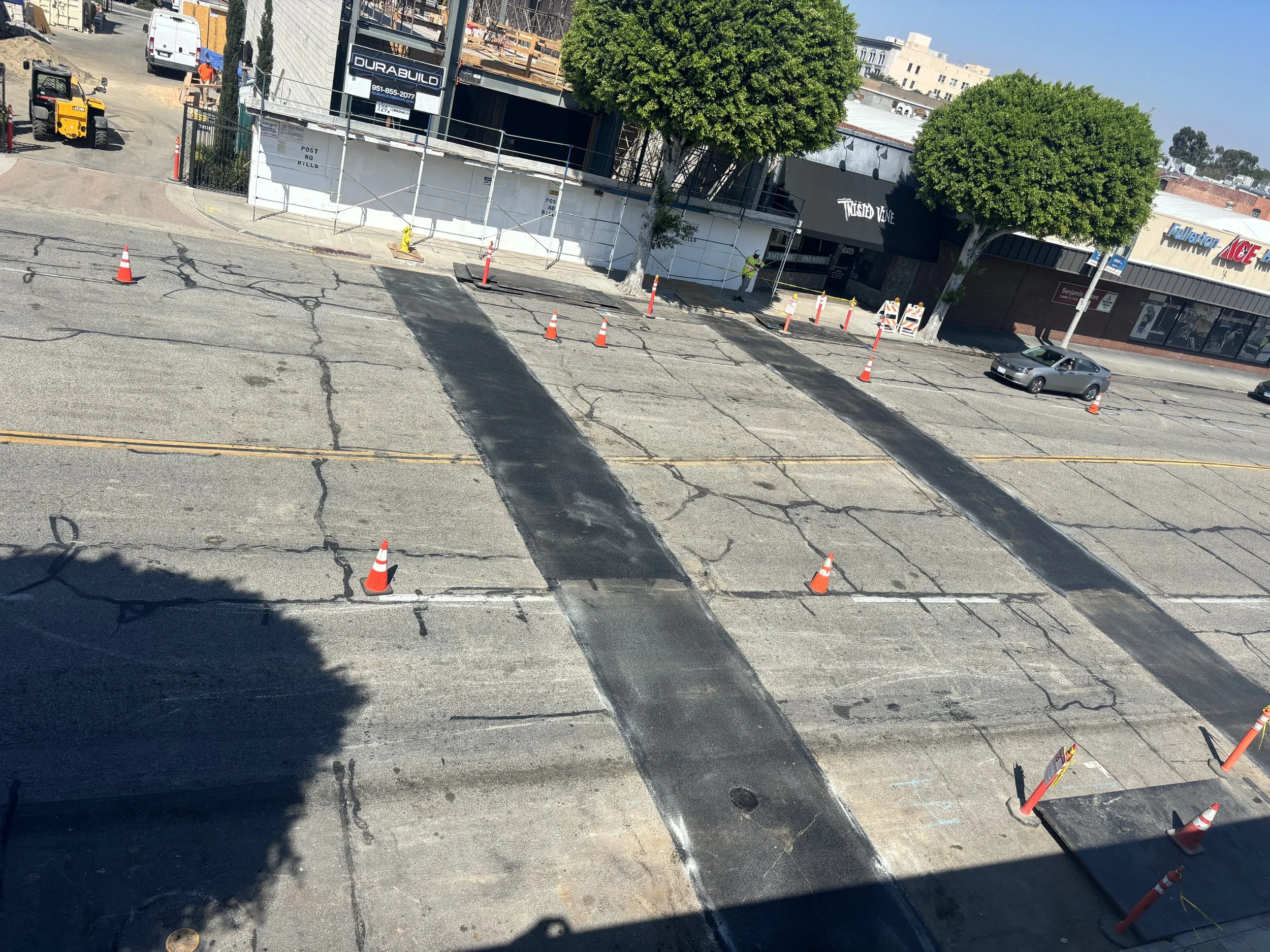 Street with sections of asphalt being repaired, marked by orange traffic cones, with two newly paved lanes and surrounding storefronts and trees.