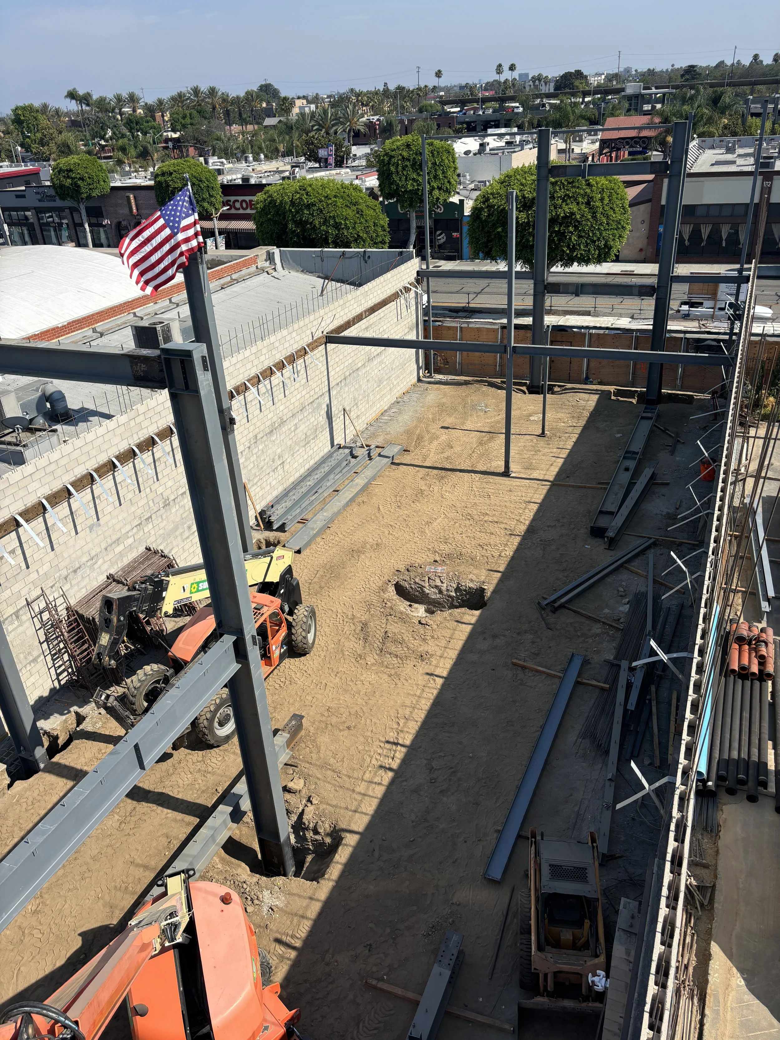 Construction site with a partially built structure, steel beams, construction vehicles, and a hole in the ground, located in an urban area with trees and buildings in the background.