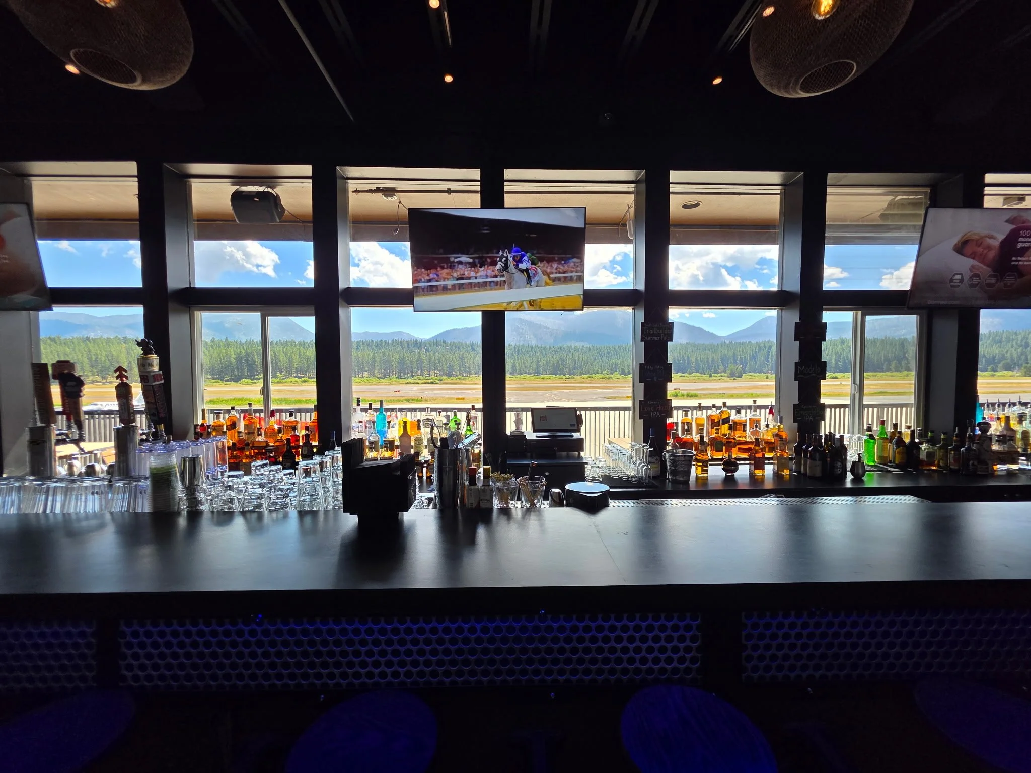 View of a bar with bottles of liquor and glasses, overlooking an airfield and mountains through large windows, with a television screen displaying horse racing.