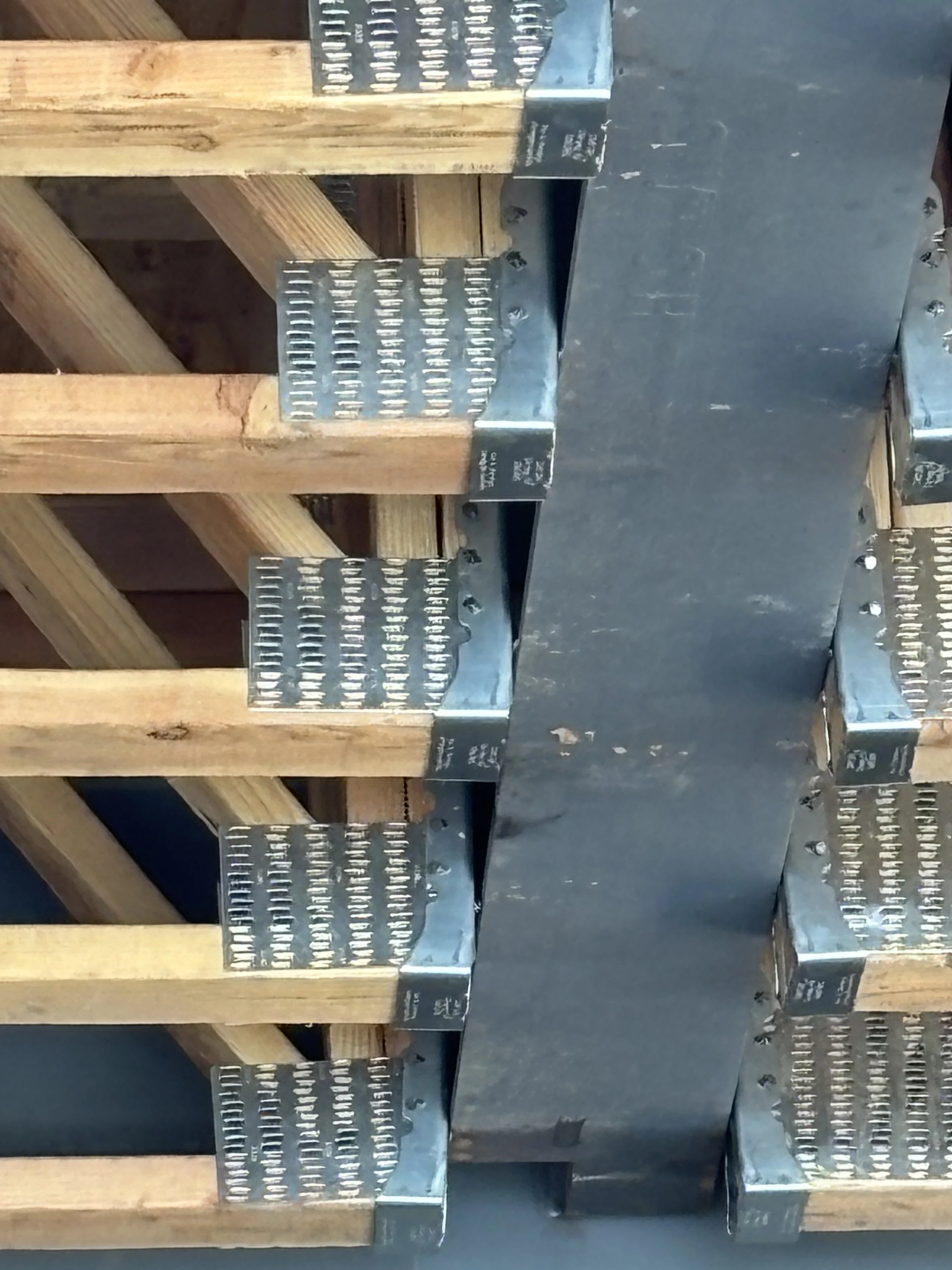 View of a construction site showing wooden framing and metal brackets attached to the wood, with a concrete surface in the background.