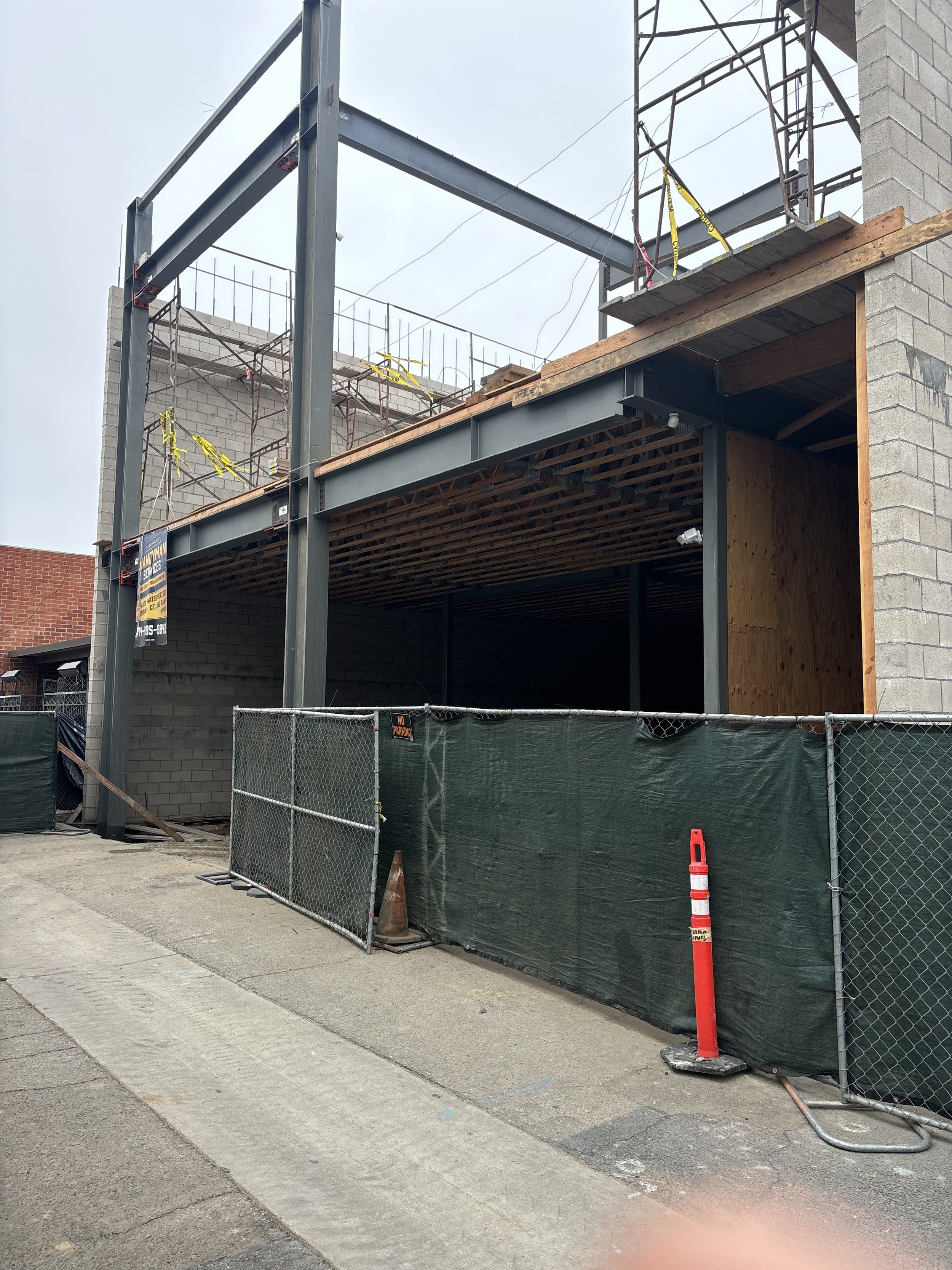 Construction site with steel framework, partially built brick wall, wooden platform, and safety barriers on sidewalk.