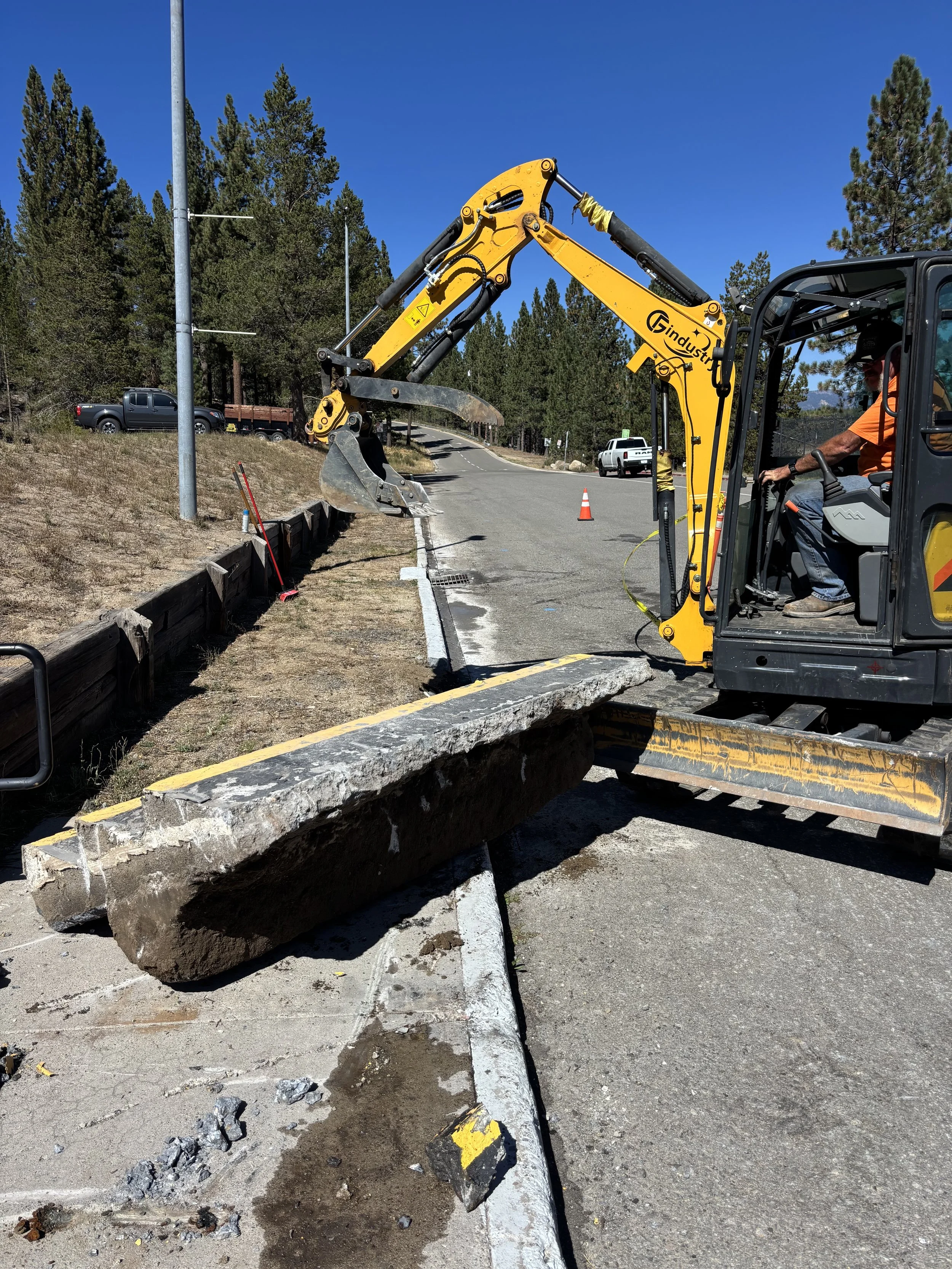 Construction worker operating a small yellow excavator removing a large concrete curb from the side of a road in a wooded area on a sunny day.