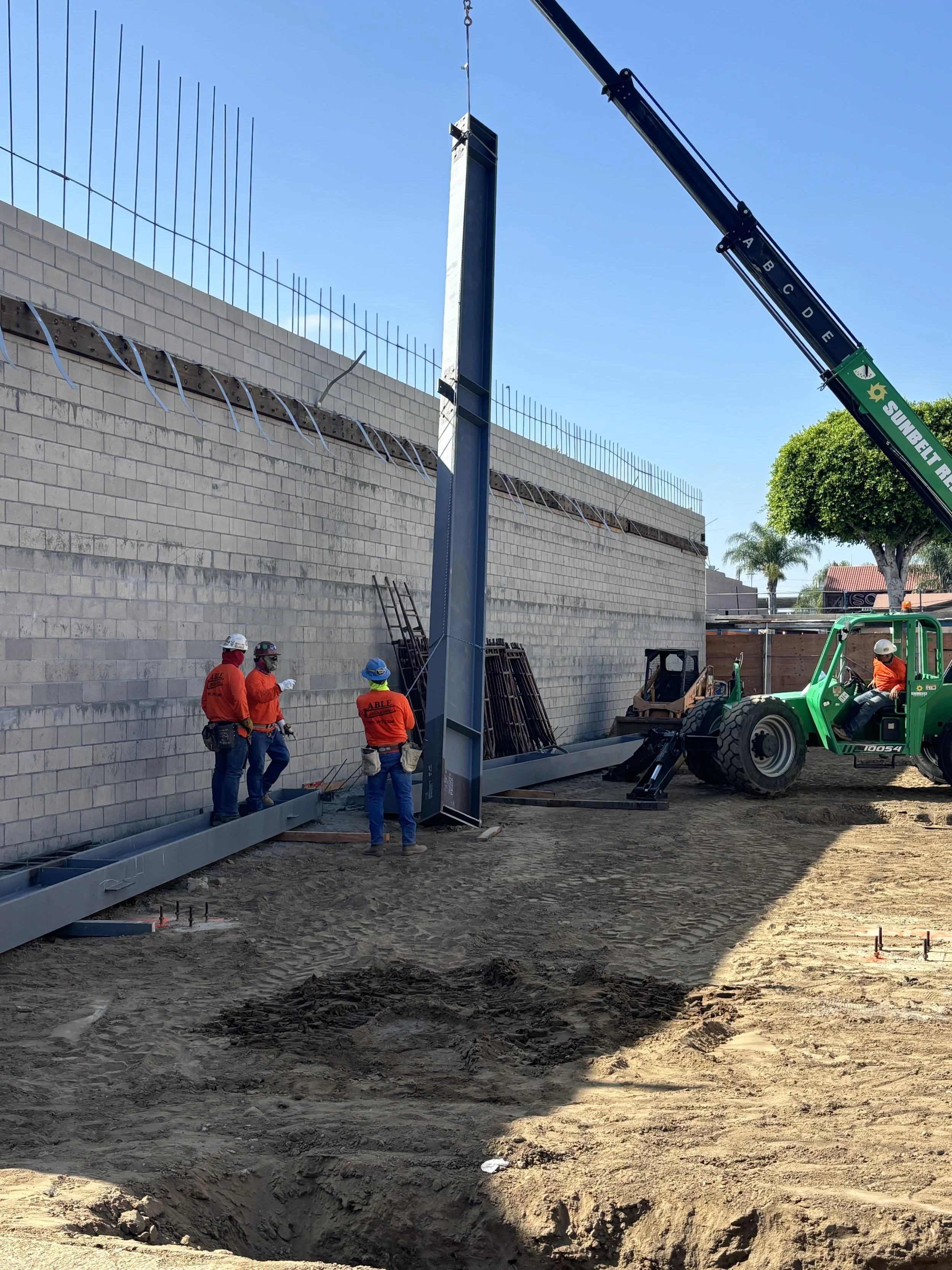 Construction workers in orange safety shirts and helmets operate a crane while assembling a tall, gray structure at a construction site adjacent to a brick wall with rebar on top. The ground is dirt, and there is a shadow cast across part of the site