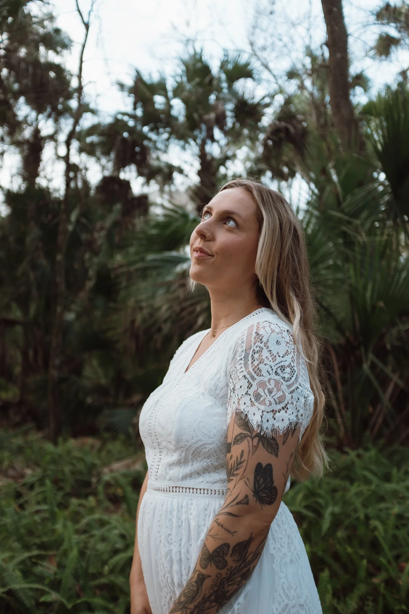 A woman with long blonde hair and tattoos on her left arm, wearing a white lace dress, standing outdoors in a forested area with tall trees and green foliage, looking upward.