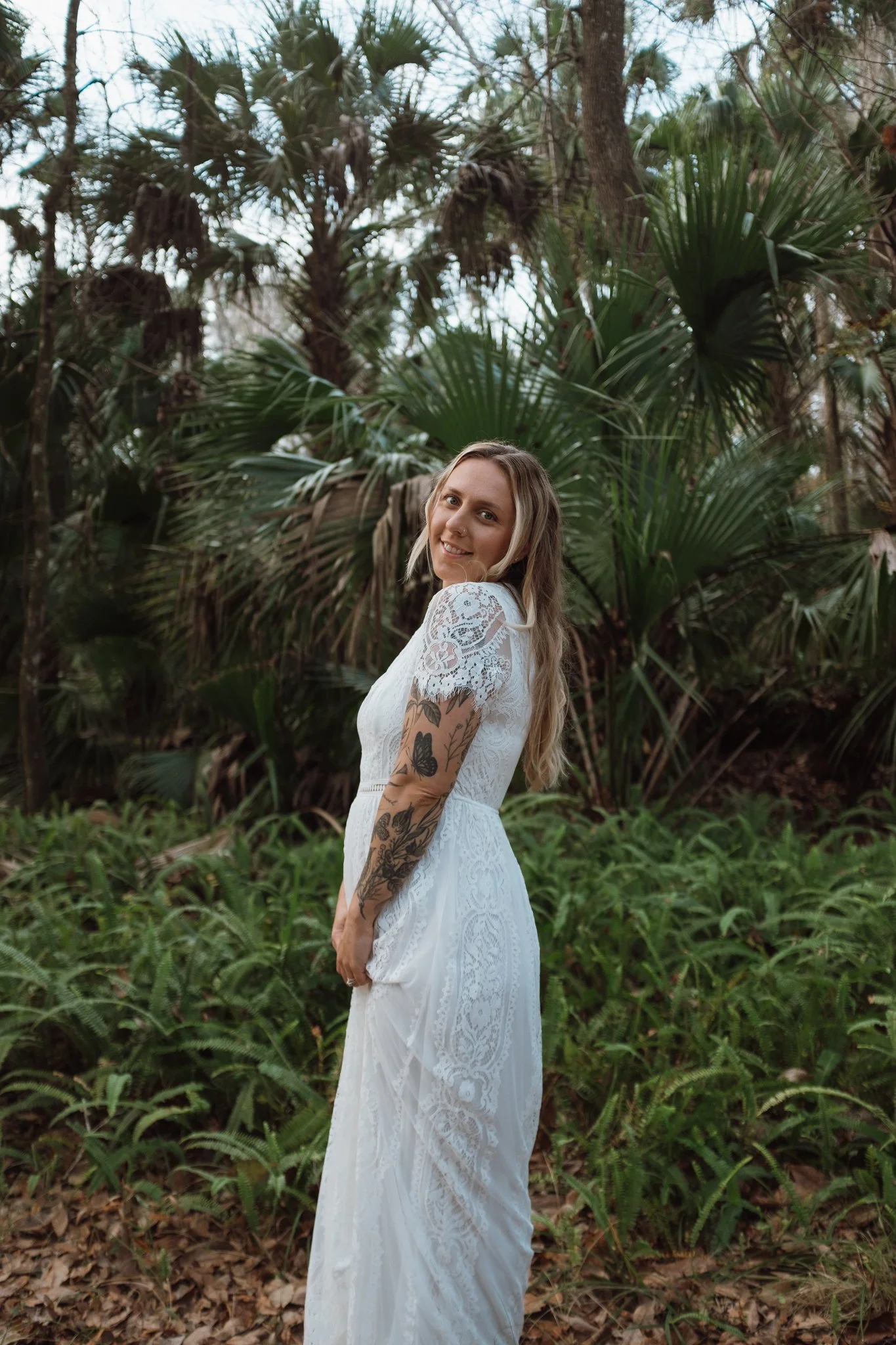 A woman with long blonde hair and tattoos on her arm wearing a white lace dress standing in a lush, green forest with various trees and plants.