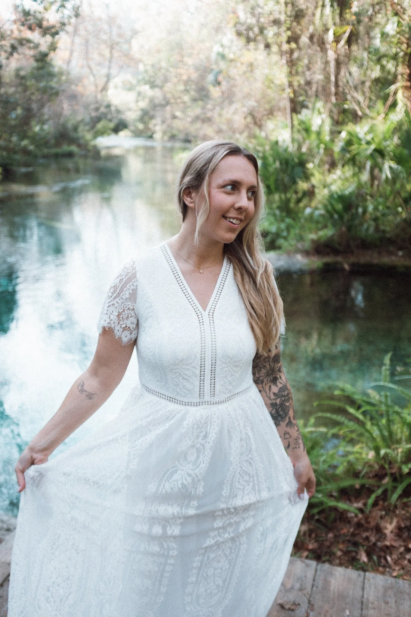 A woman in a white dress standing outdoors next to a river or creek surrounded by trees.