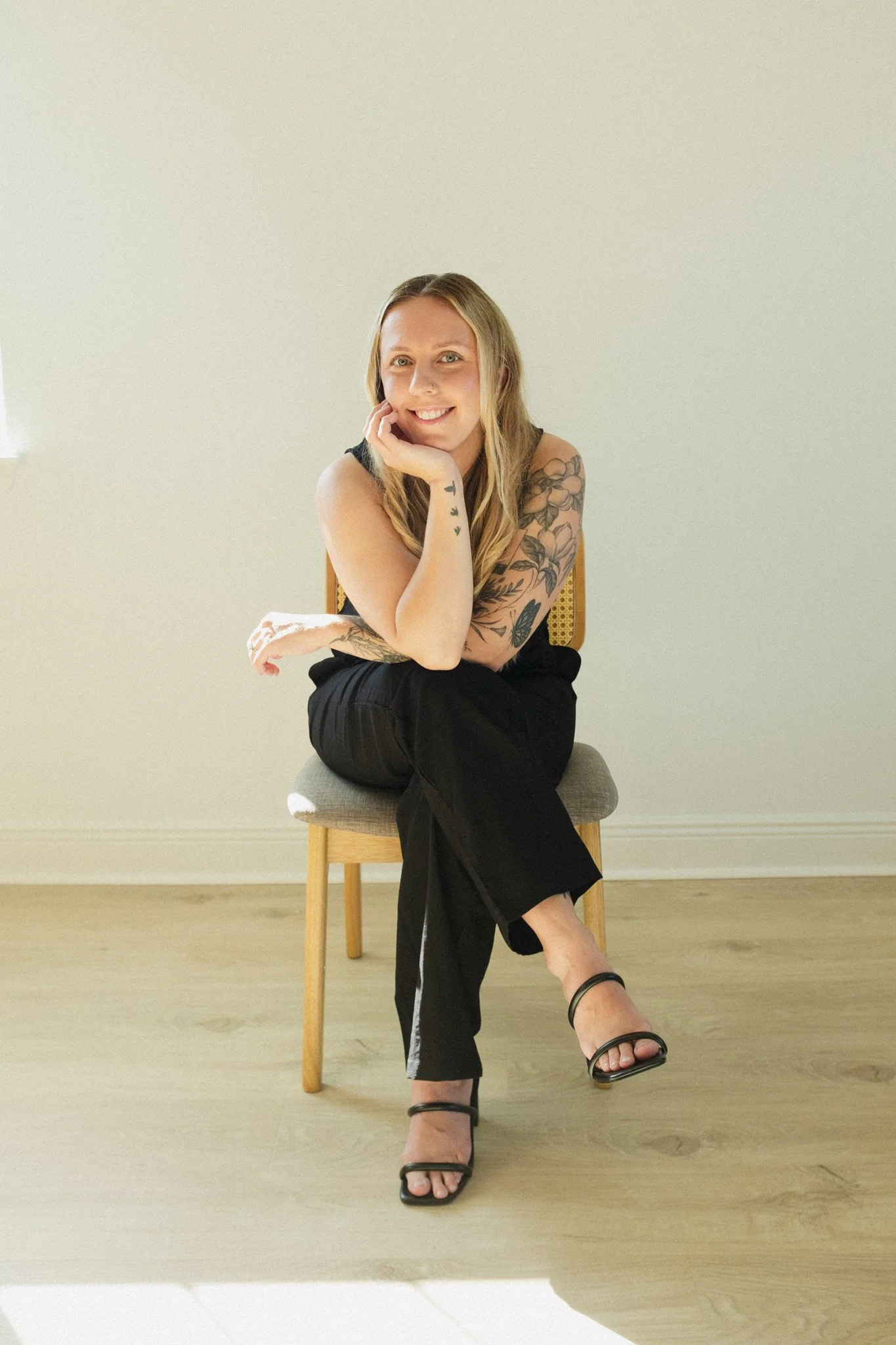 A woman sitting on a wooden chair in a room with a plain wall. She has long blonde hair, tattoos on her arms, and is smiling at the camera. She wears a sleeveless top, black pants, and black high-heeled sandals.