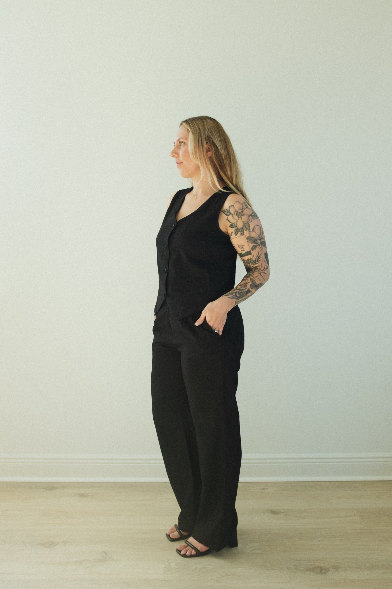 A woman with long blonde hair and tattoos on her left arm, wearing a black sleeveless vest and black pants, standing against a plain white wall.