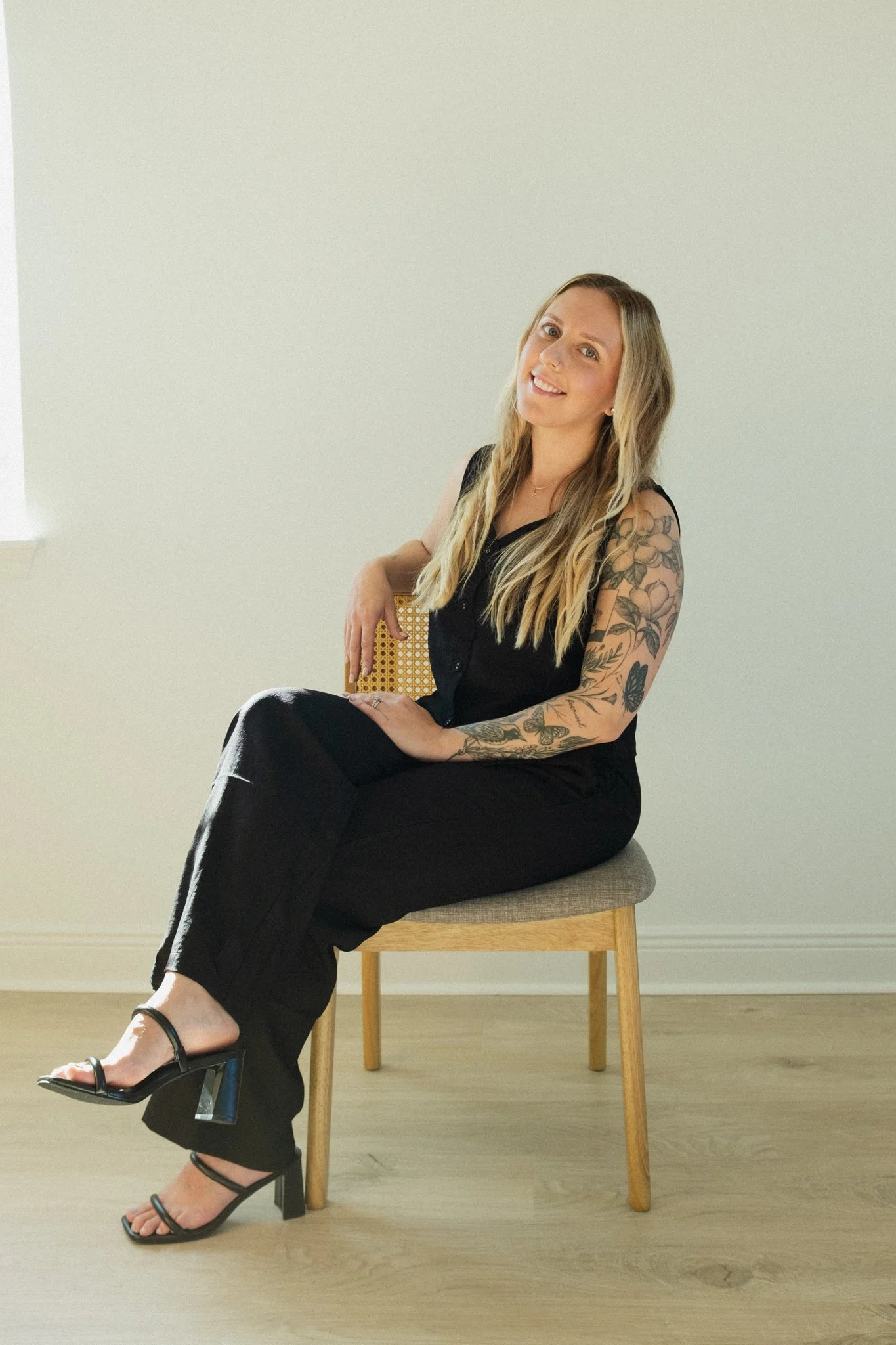 A woman with long wavy blonde hair and tattoos on her arms, wearing a black sleeveless top and black pants, sitting on a beige chair against a plain light-colored wall.