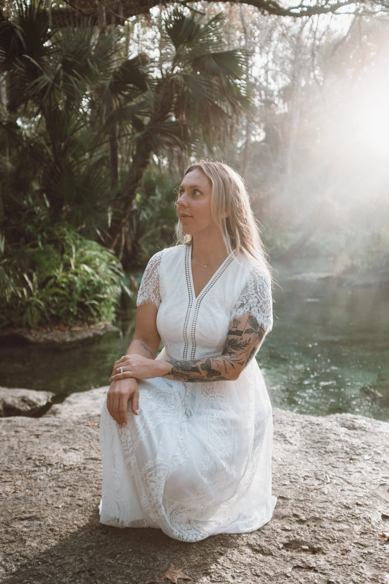 A woman with long blonde hair and tattoos on her arms, dressed in a white lace dress, kneeling on a rock near a body of water in a forest; sunlight shining through trees.
