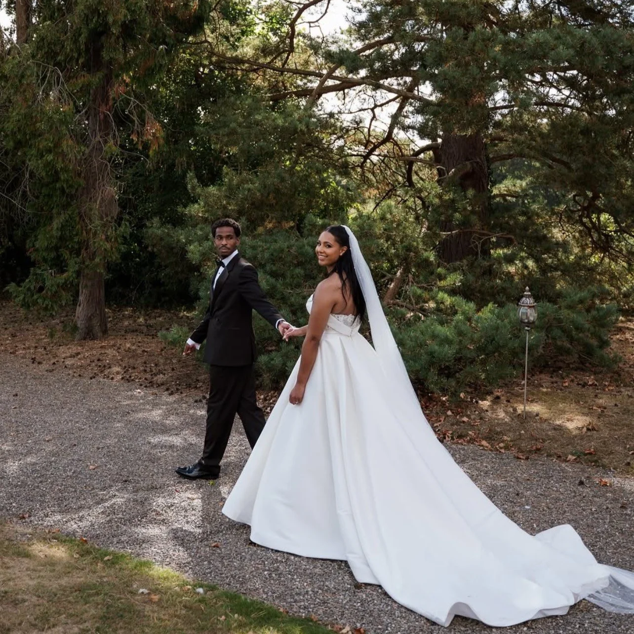 Bride and groom holding hands on a pathway outdoors, with trees and bushes in the background, during a wedding.