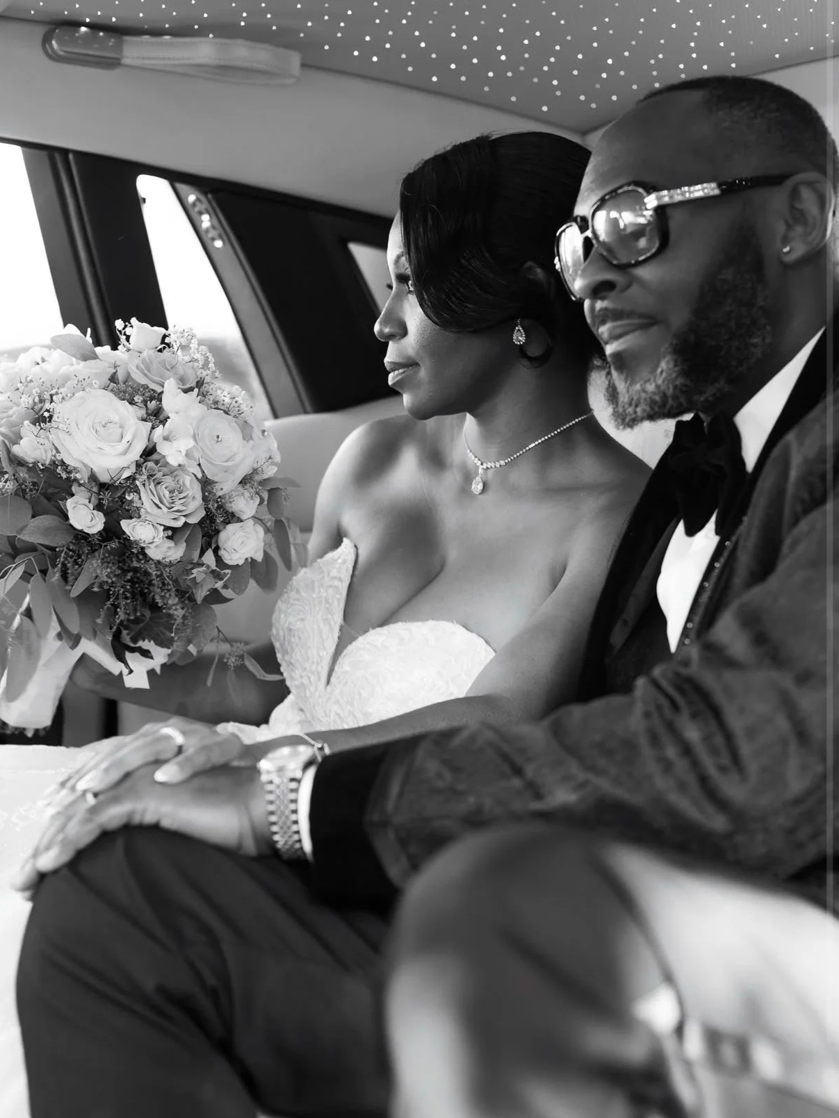 Black and white photo of a bride and groom sitting together in the back of a vehicle, with the bride holding a bouquet of roses.