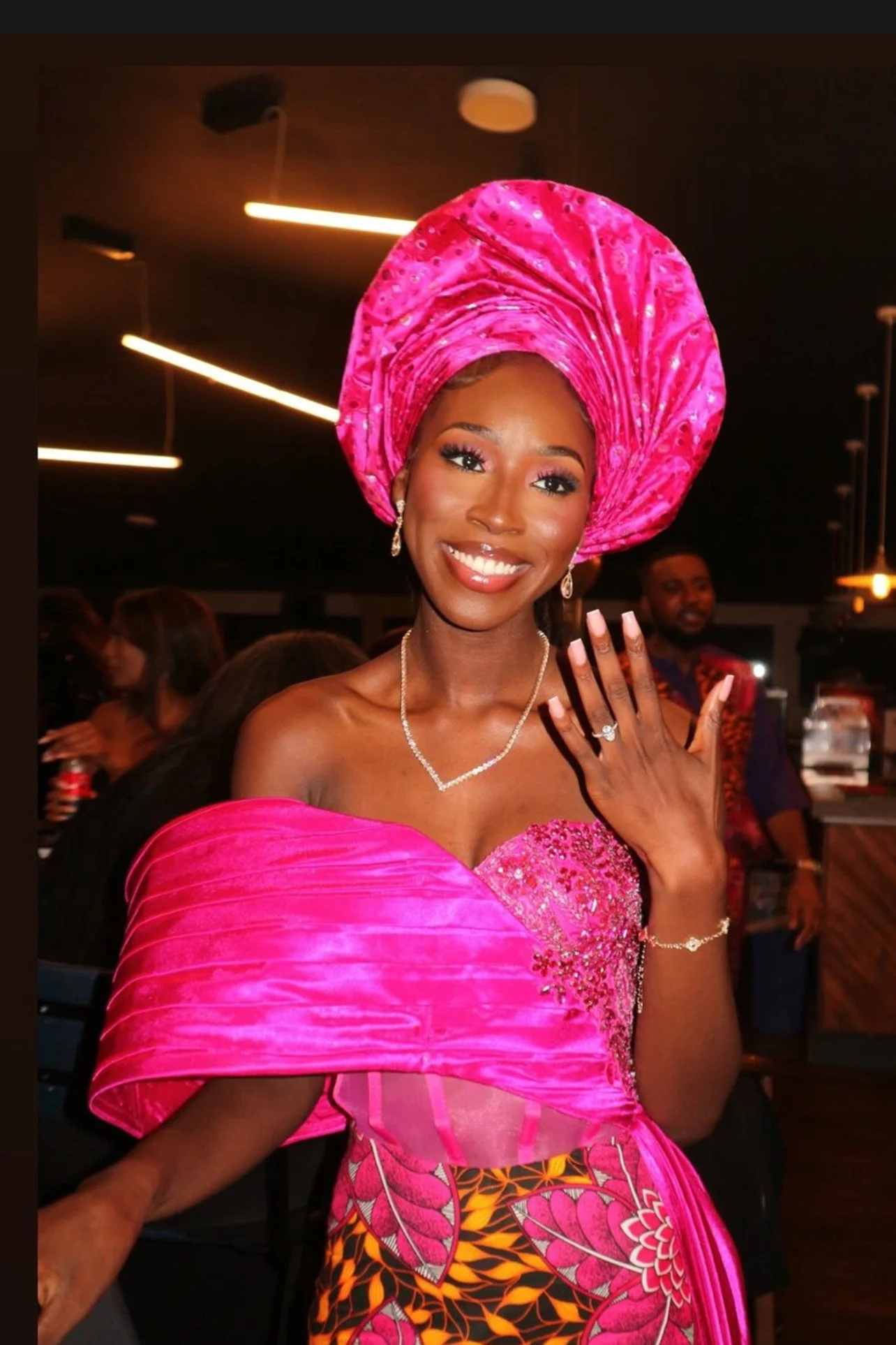 A woman dressed in vibrant traditional attire, wearing a bright pink gele headwrap and matching dress with intricate beading, smiling at an indoor event.