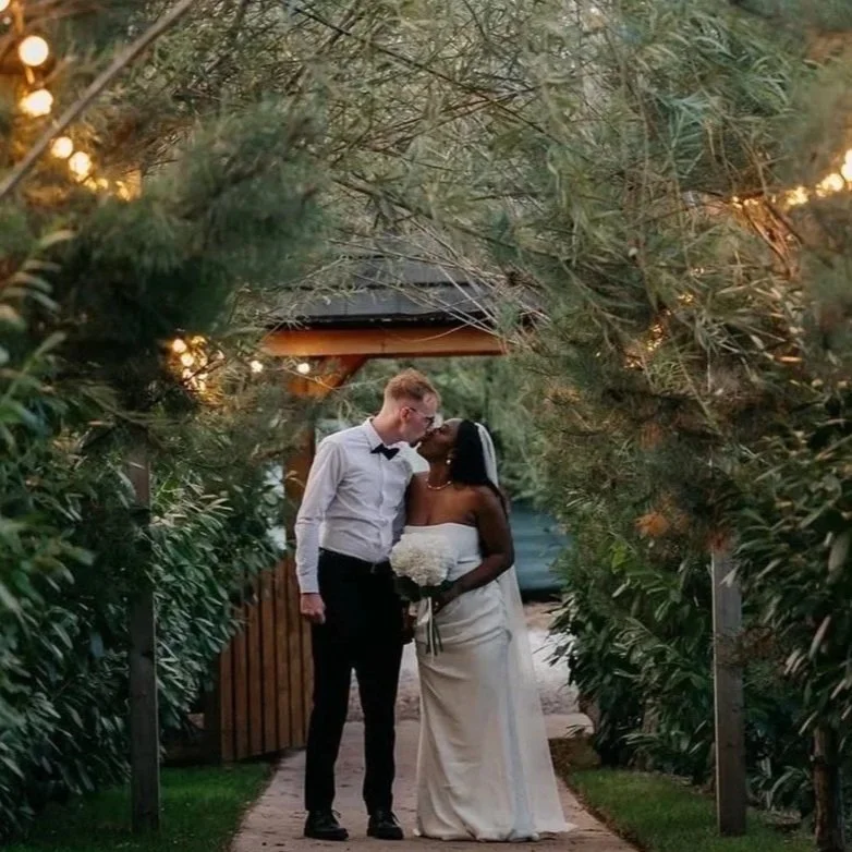 A newlywed couple sharing a kiss outdoors amid greenery and string lights, with a wooden pavilion backdrop.
