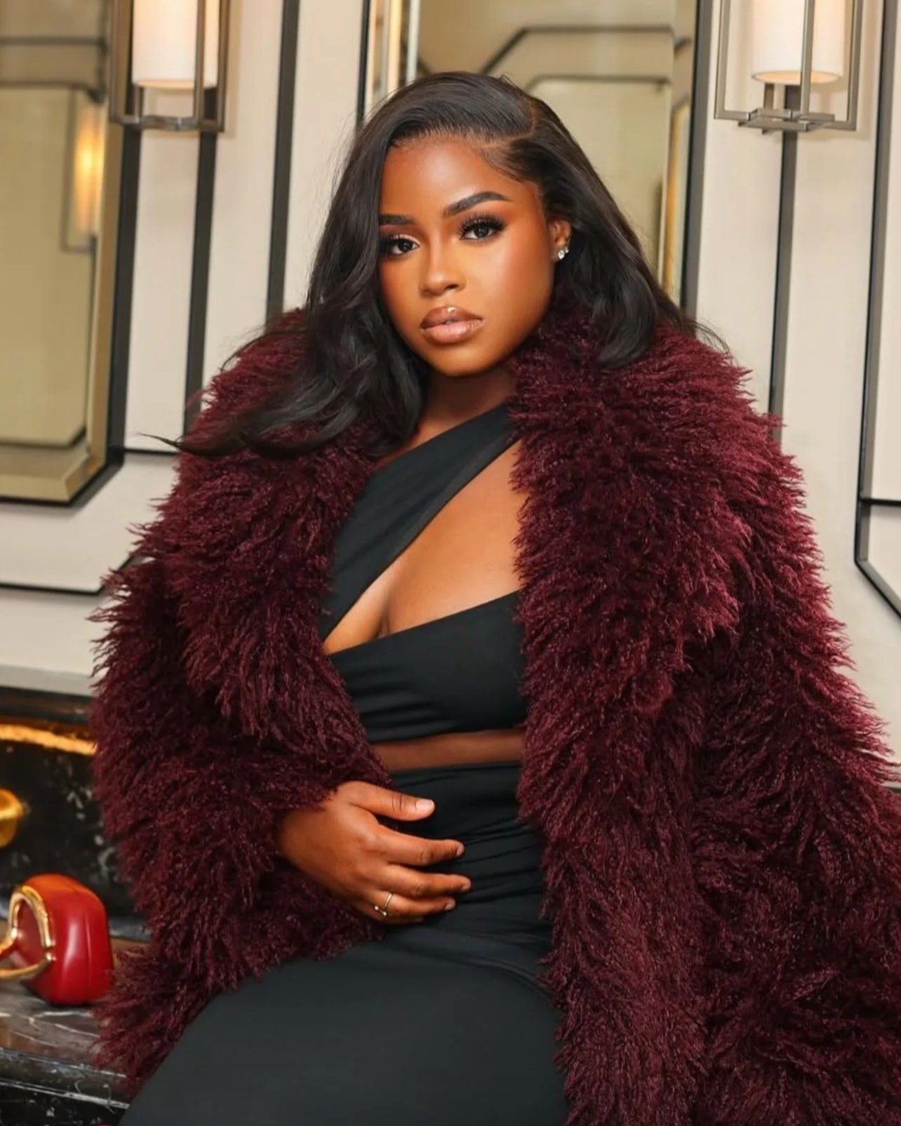 A woman with dark hair, wearing a black dress with a deep neckline, a maroon furry coat, and sparkling earrings, sitting indoors next to a marble table with a red handbag on it.