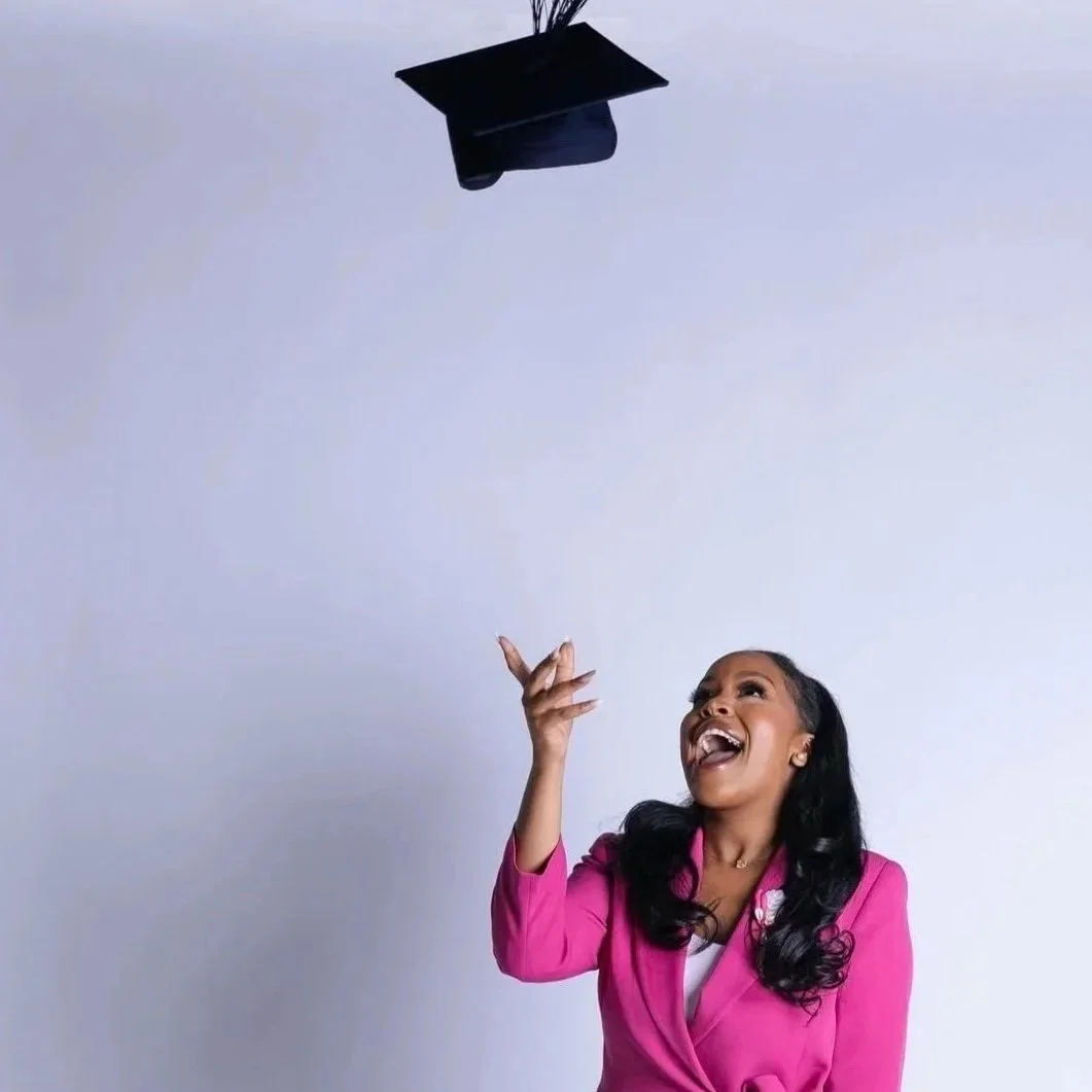 Woman in a bright pink suit celebrating graduation by tossing a black graduation cap into the air, holding a rolled-up diploma.