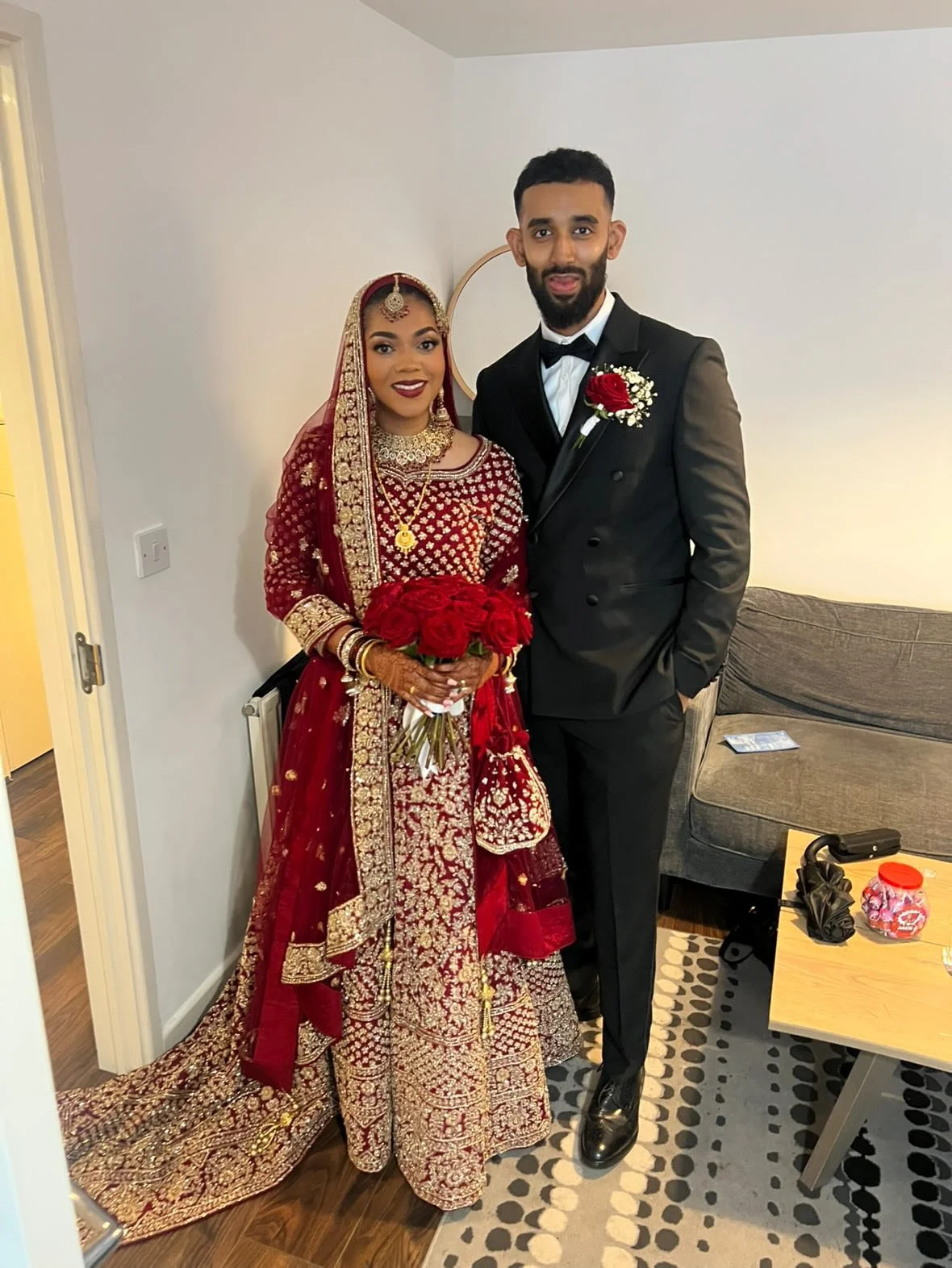 A bride and groom in wedding attire standing together indoors, with the bride holding a bouquet of red roses.