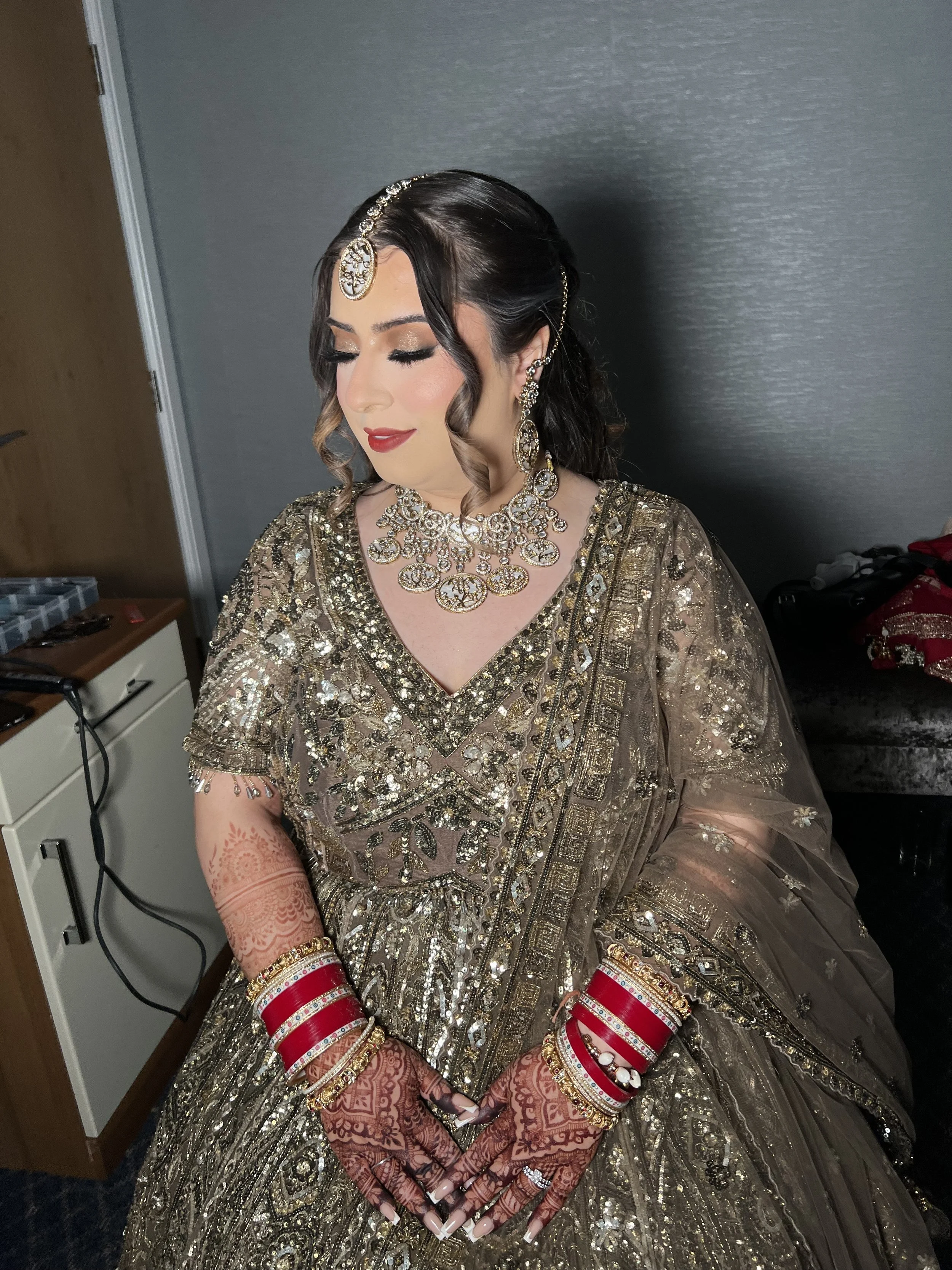 A woman dressed in traditional Indian bridal attire with ornate gold and silver embroidery, adorned with jewelry including earrings, necklaces, bangles, and a headpiece, with henna designs on her hands.