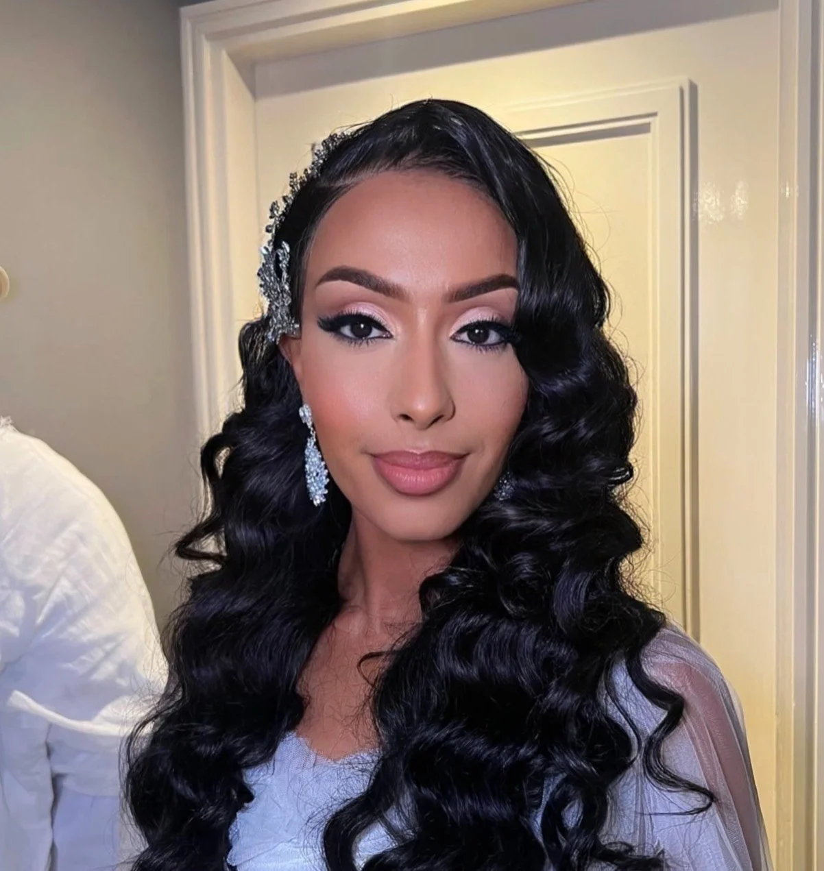 A woman with long, dark, curly hair is looking at the camera and smiling softly. She is wearing glamorous makeup, large earrings, and a decorative hair accessory with rhinestones.