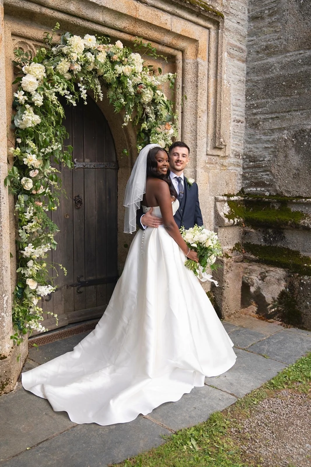 A bride and groom stand close together outside a stone building decorated with a floral arch. The bride wears a white wedding gown with a veil and holds a bouquet, and the groom wears a dark suit with a white shirt and tie.